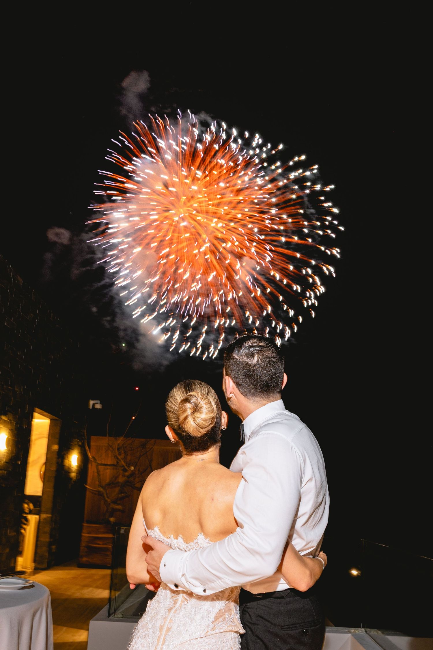 Couple embracing while watching vibrant orange fireworks light up the night sky at a wedding celebration.
