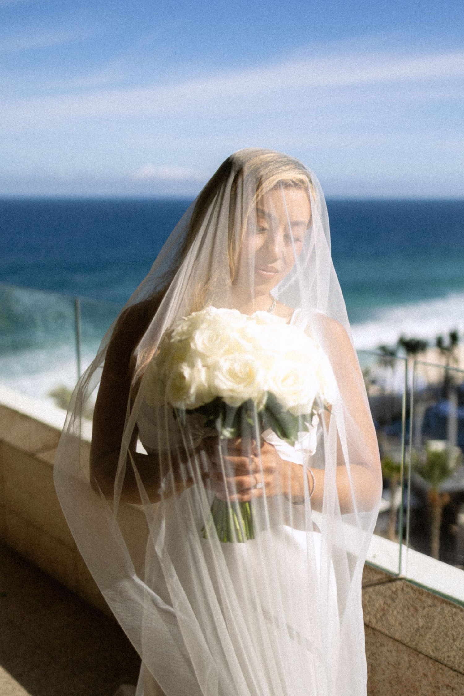 Sunlit bridal portraits through delicate white veil with ocean vista in background.