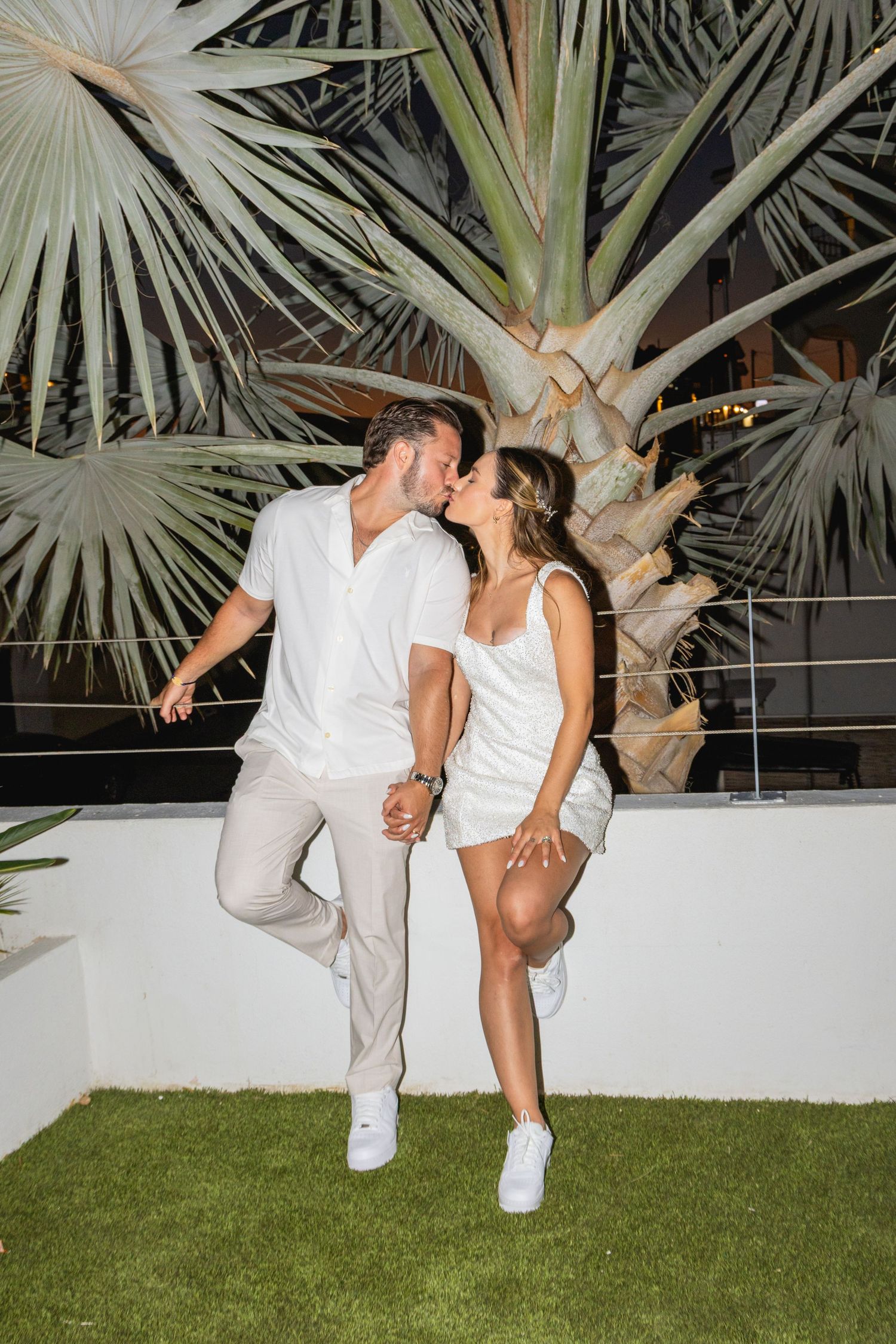 A couple in all-white outfits dances playfully on grass with palm trees in the background.