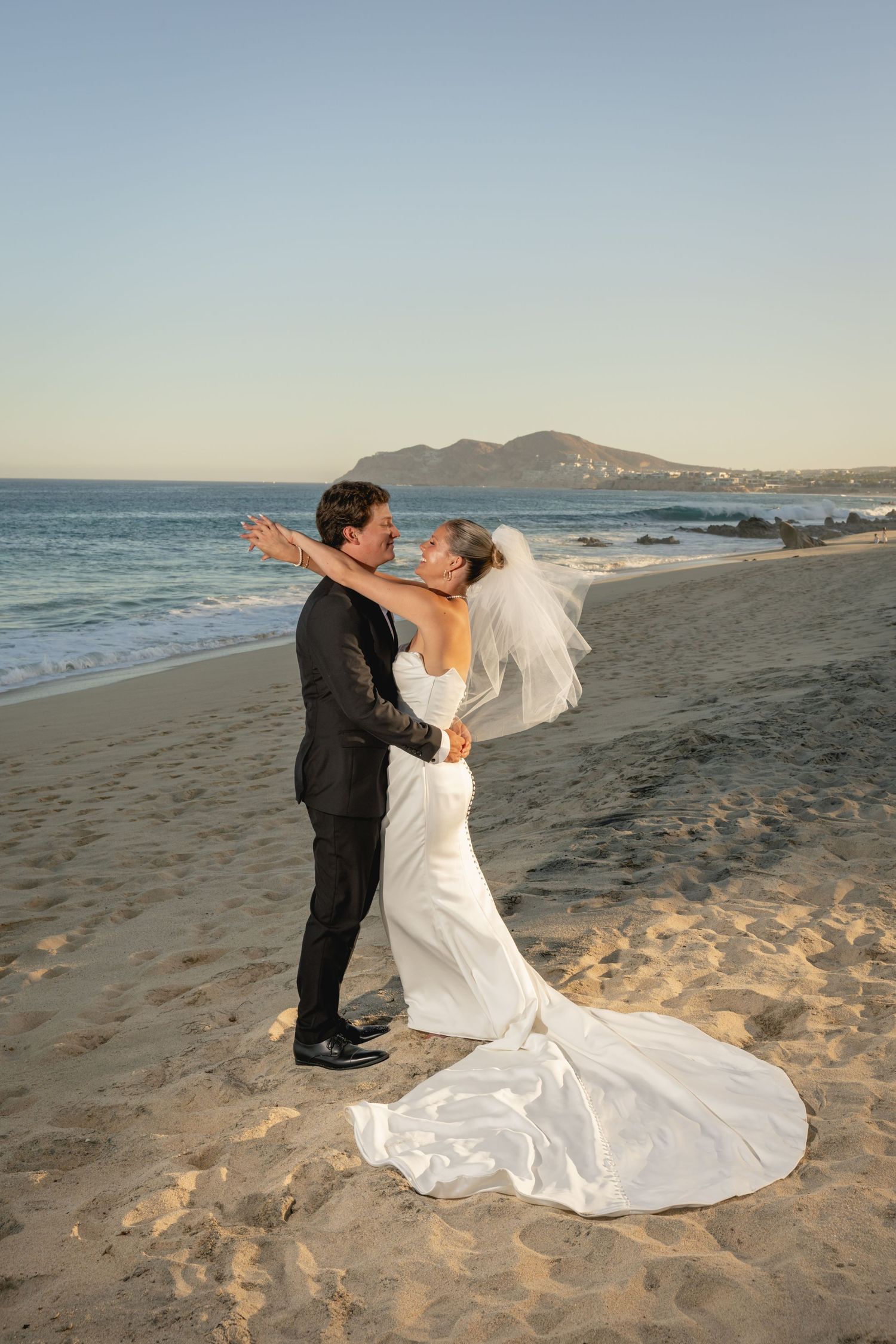 Bride and groom embrace on a sandy beach at sunset, with ocean waves and a mountain in the background.
