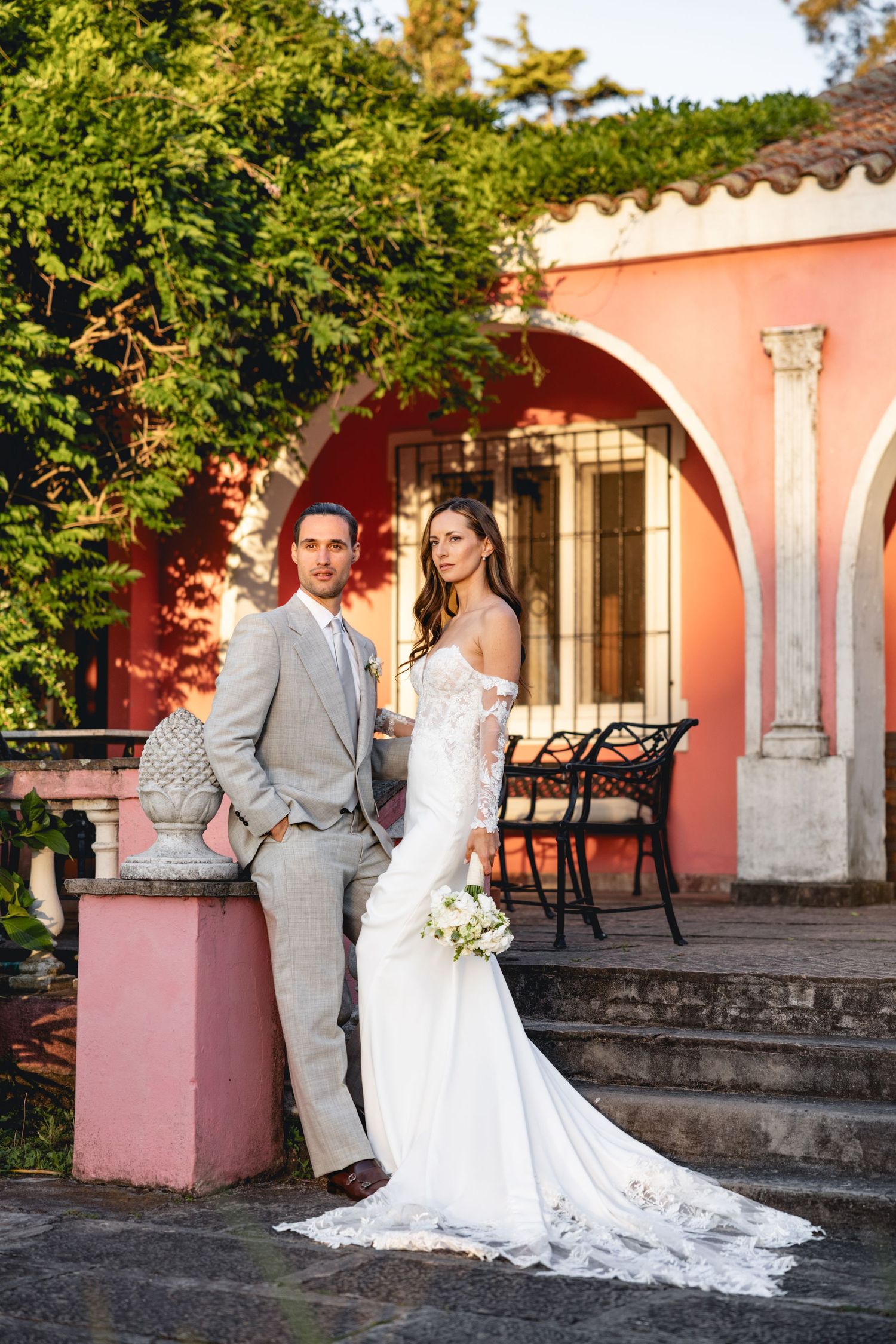 Wedding couple poses along pink Mediterranean-style architecture with trailing lace dress.