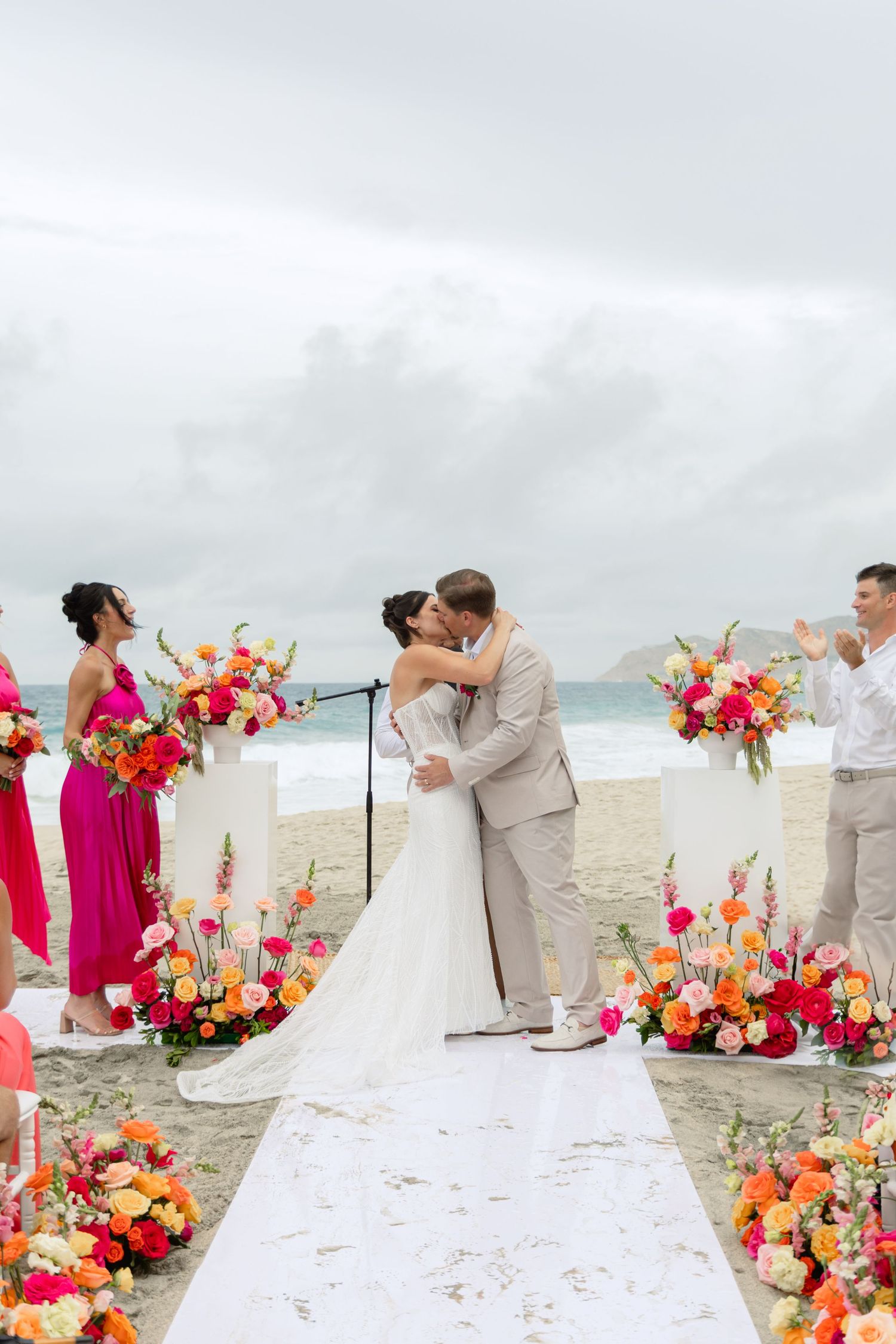 Beach wedding ceremony with pink and orange floral decorations on white aisle runner.