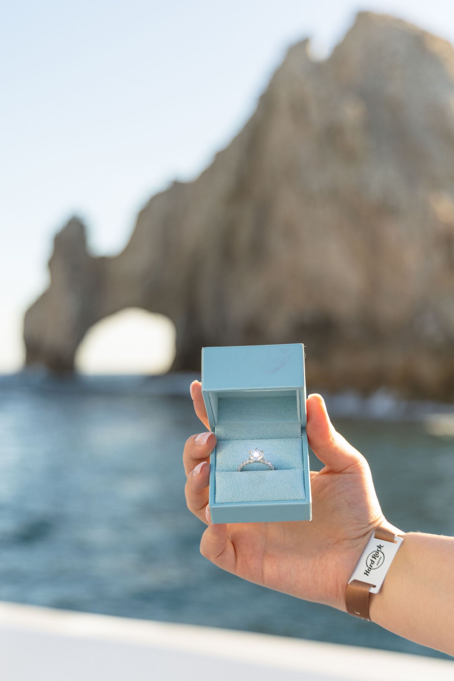 Hands holding a small turquoise jewelry box against a blurred coastal background with rock formations.