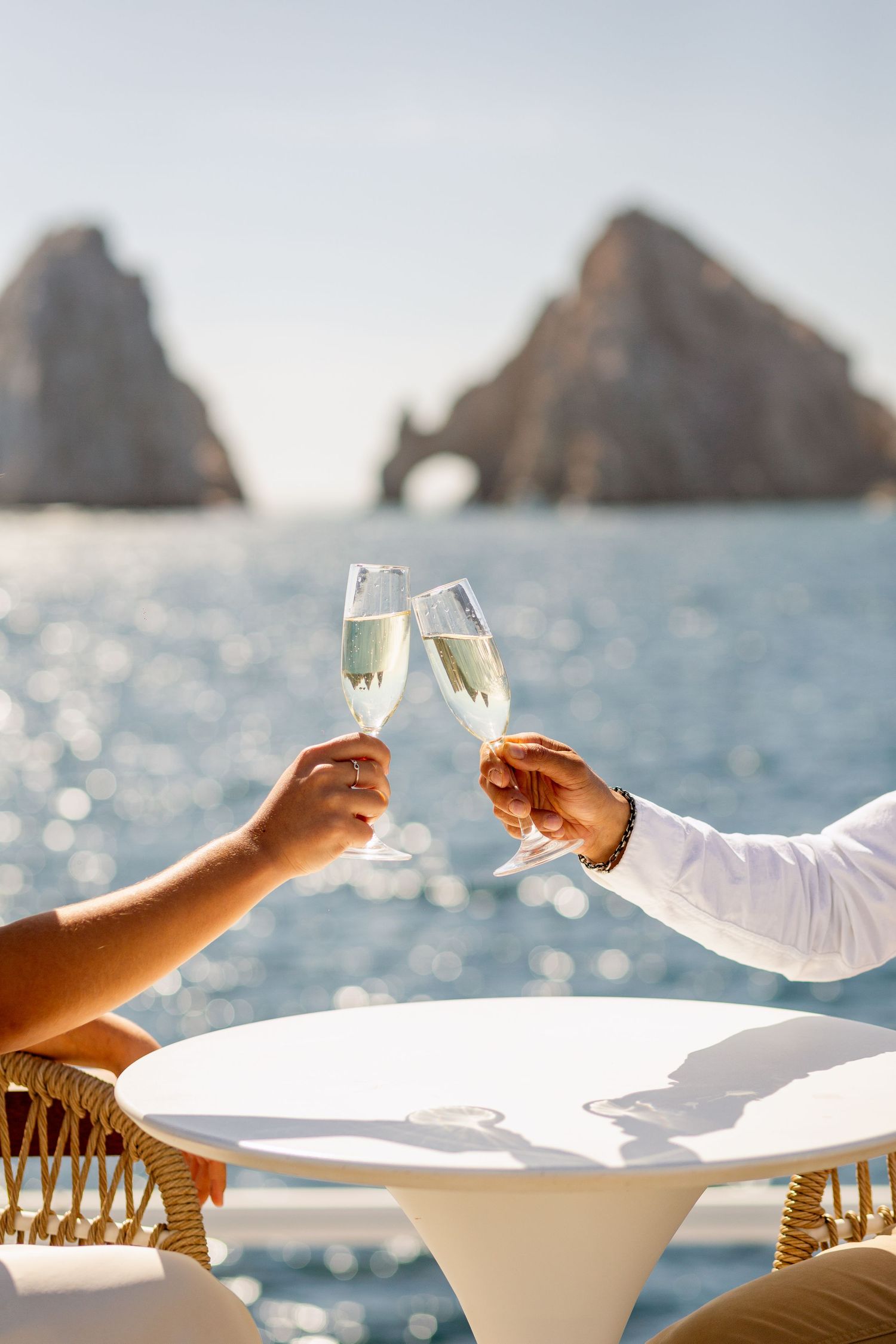 Two people toasting with champagne glasses against a scenic ocean backdrop with rocky formations.