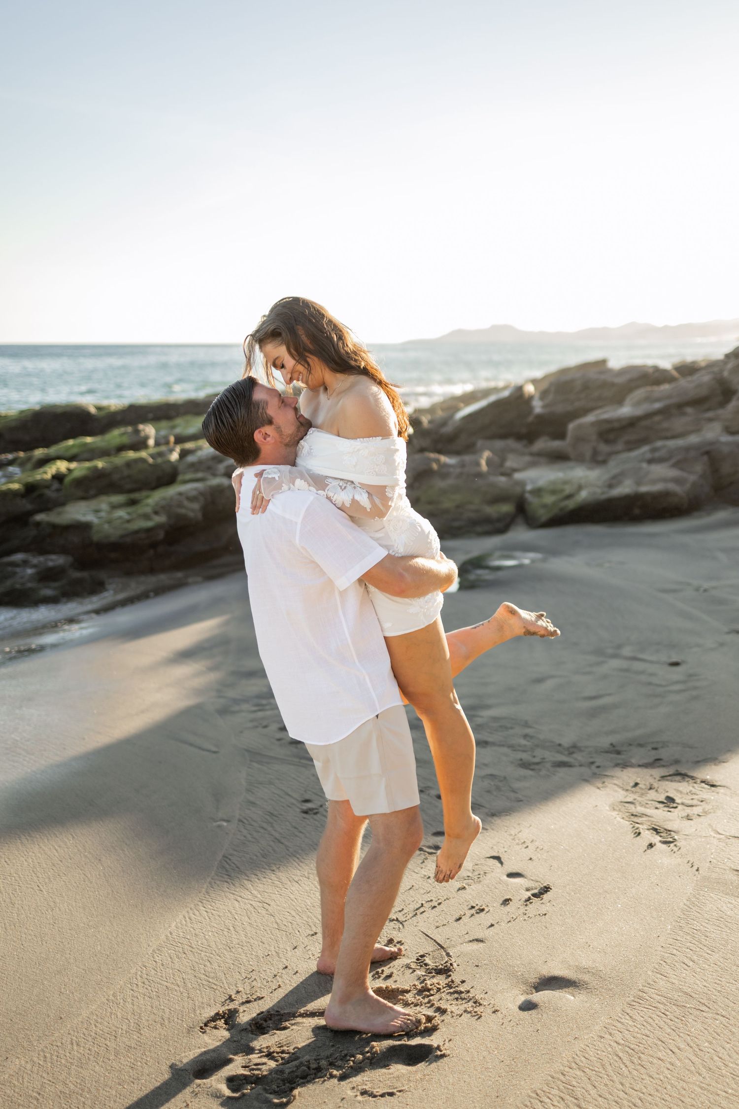 Playful couple in white clothing dancing on sandy beach near rocky coastline.