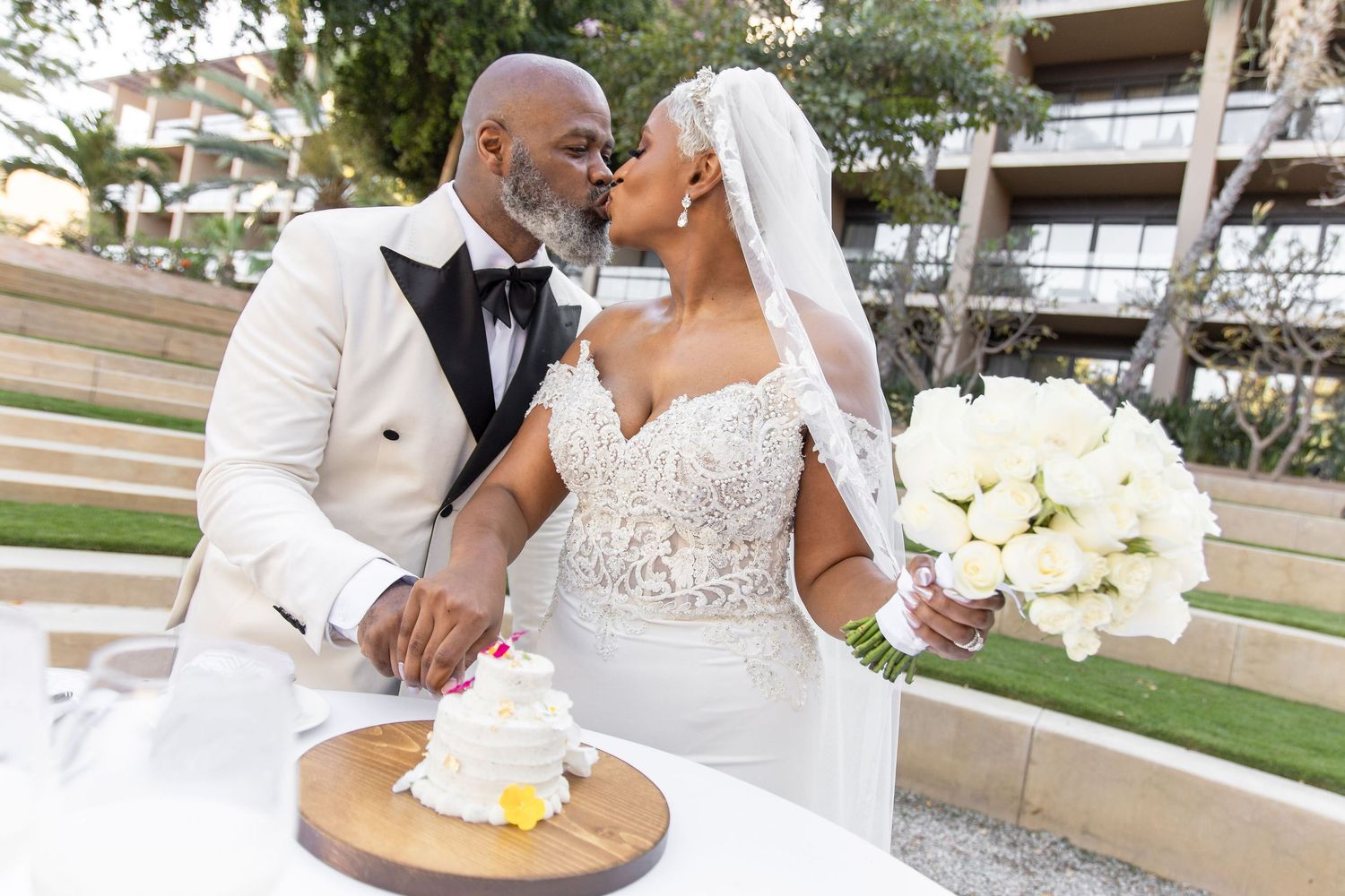 Wedding couple sharing cake cutting moment at outdoor reception with white floral bouquet.