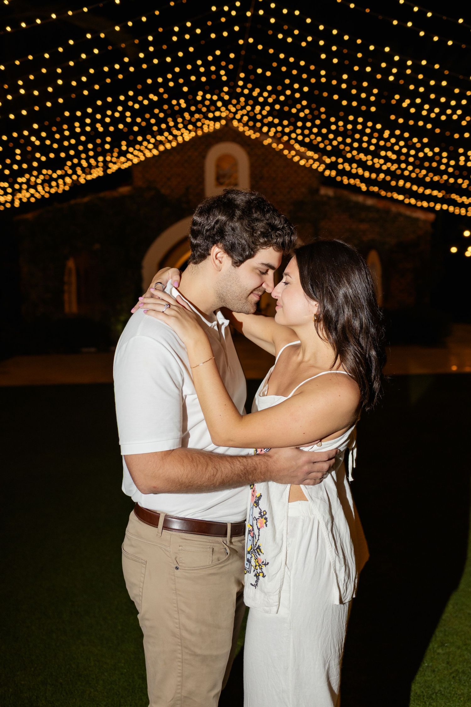 A couple shares a romantic dance under twinkling string lights at an outdoor evening celebration.