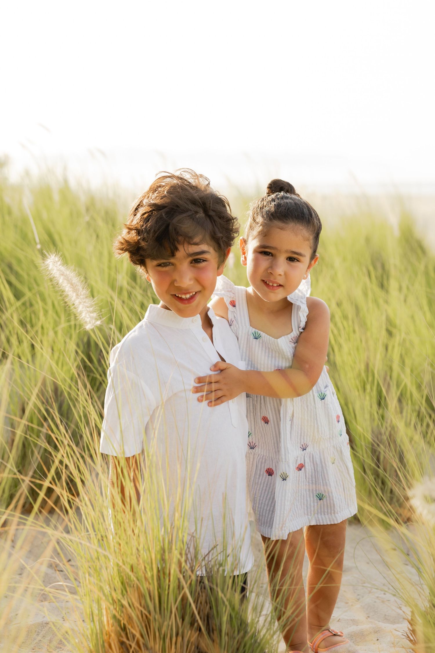 Two young children in white outfits stand among tall beach grass on sandy dunes, smiling at the camera.