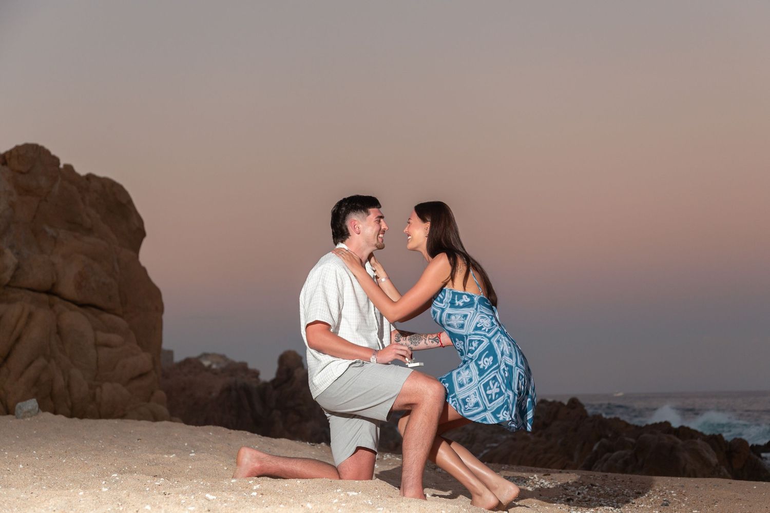 A couple shares a romantic moment on a beach at sunset with rocky cliffs in the background.