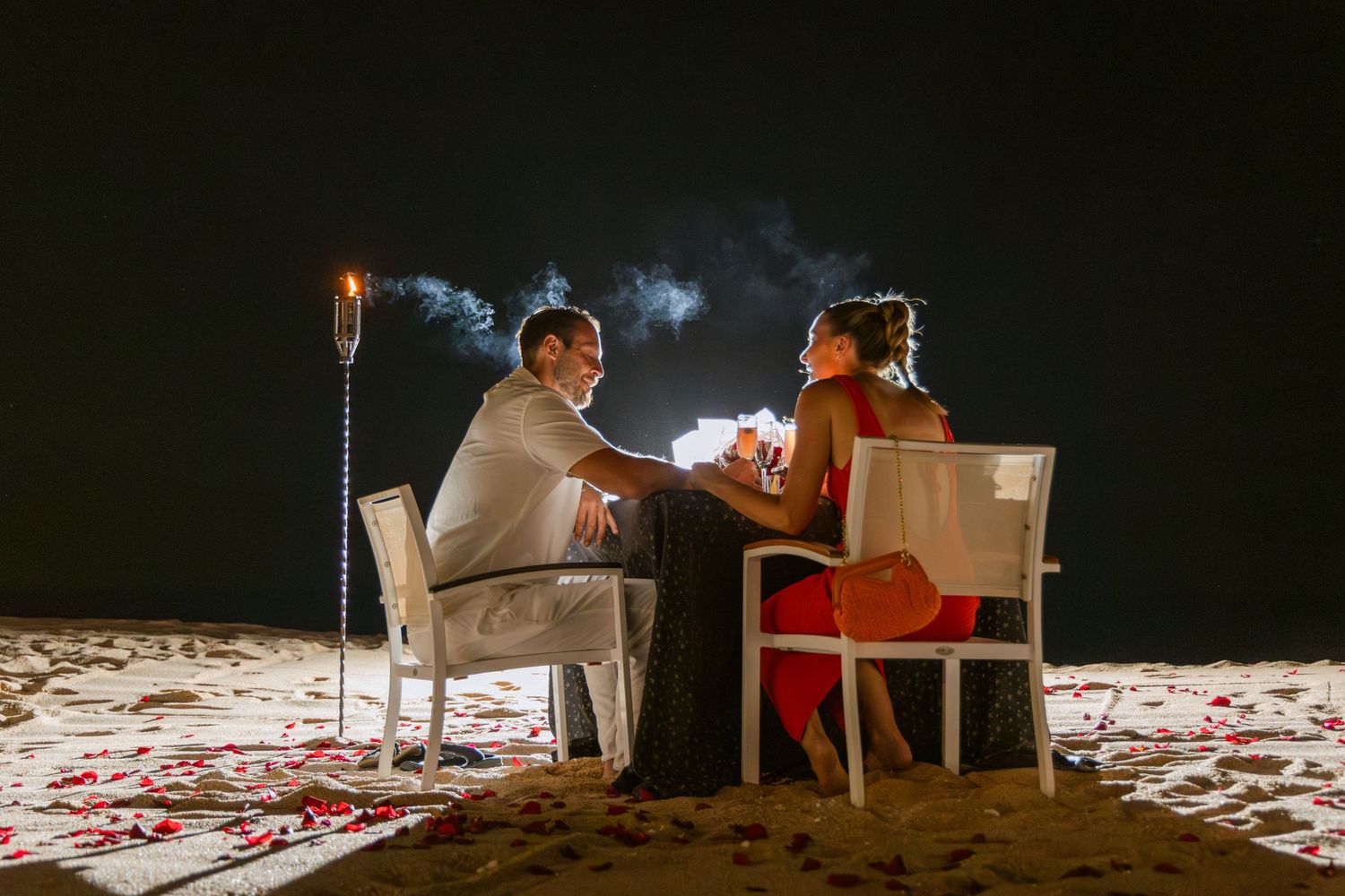 Two people sit on white chairs on a beach at night surrounded by rose petals and candlelight.