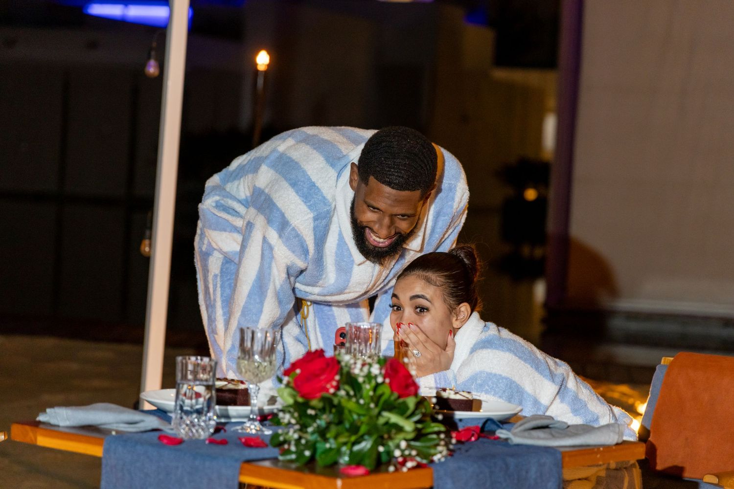 Two people in light blue outfits share a moment at an elegantly set dinner table with floral centerpiece at night.