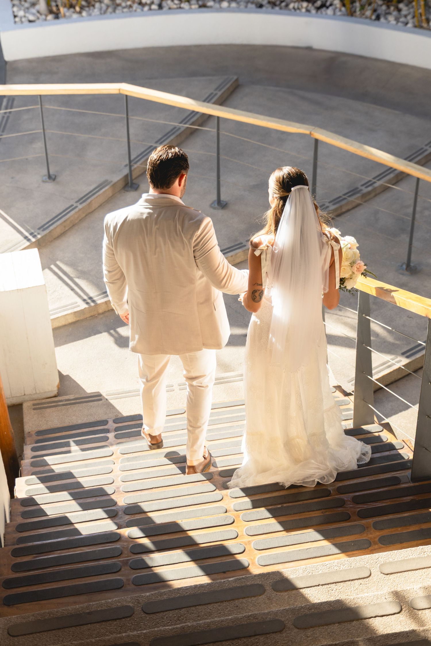 Newlyweds descending sunlit staircase holding hands in light colored attire.