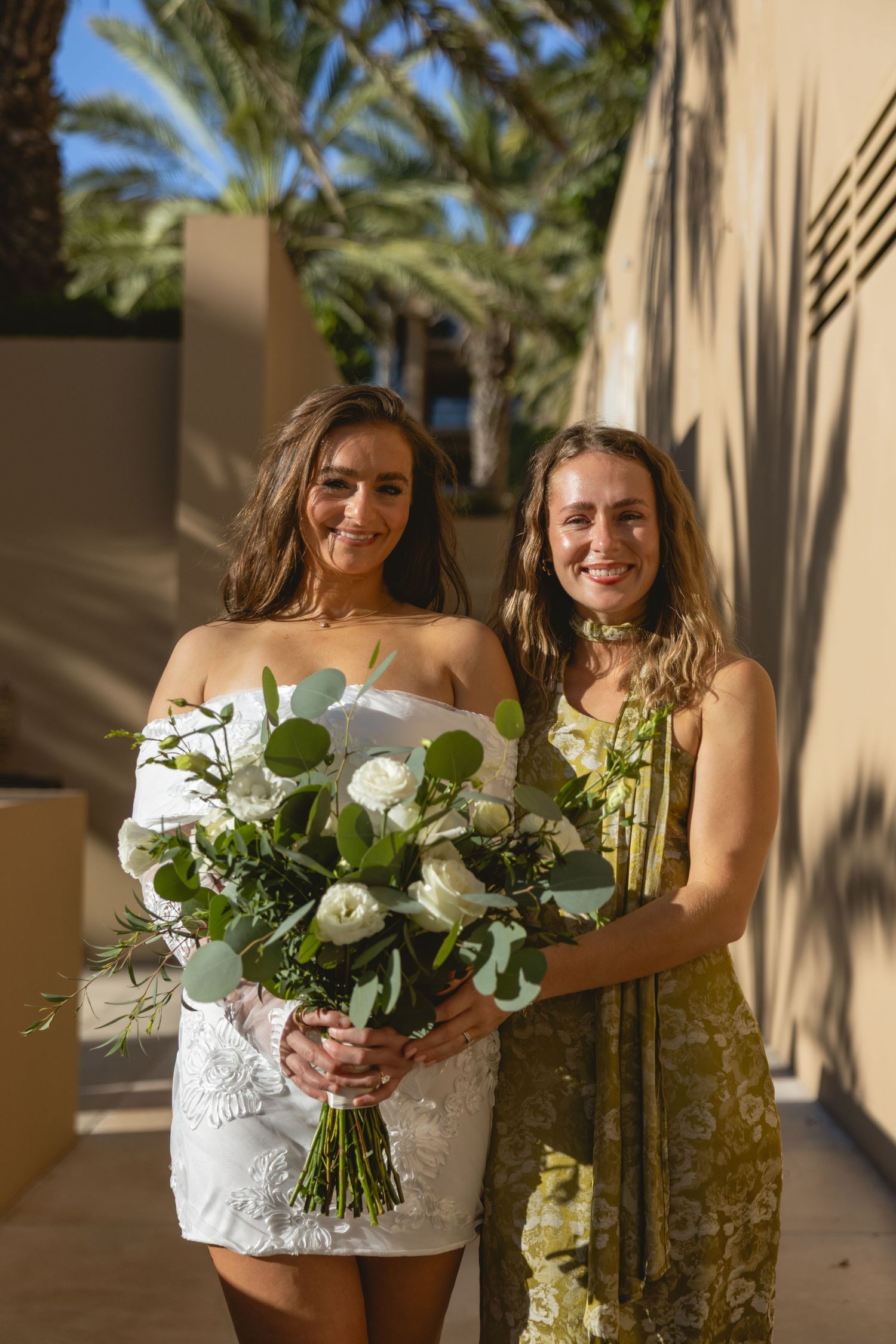 Two people pose with white floral bouquets on a palm-lined walkway during a wedding.