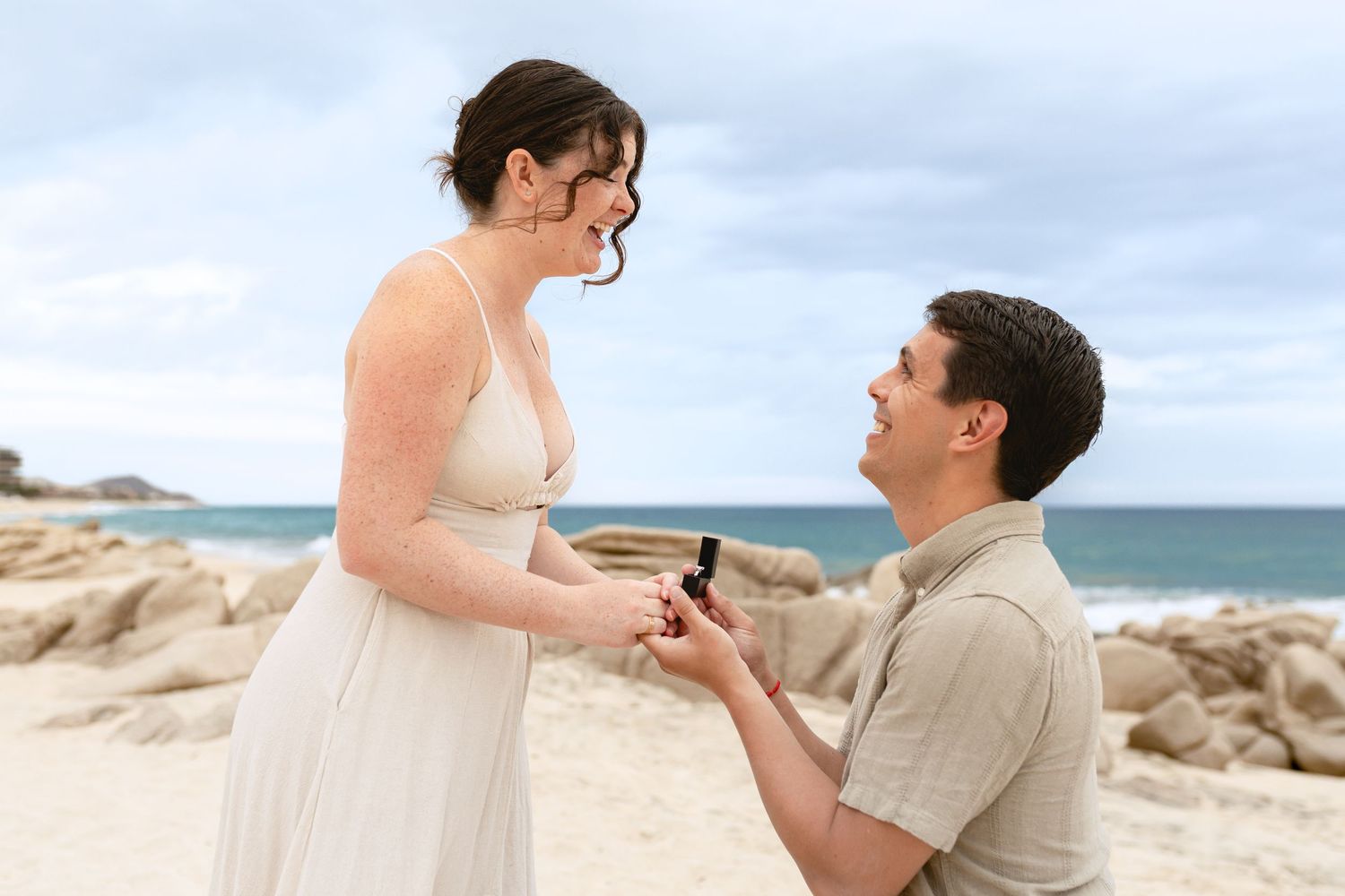 A couple shares a romantic moment during a beach proposal with ocean waves and rocky coastline in the background.