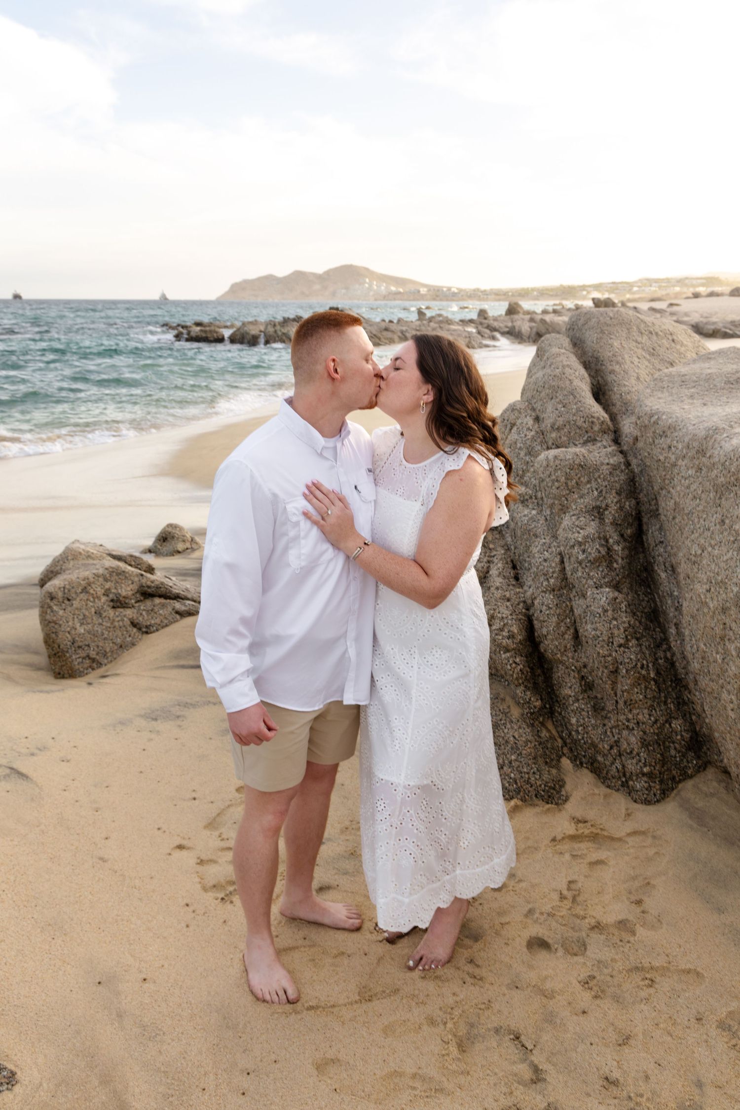 Romantic beach scene with couple in white outfits sharing tender moments by coastal rocks at sunset.