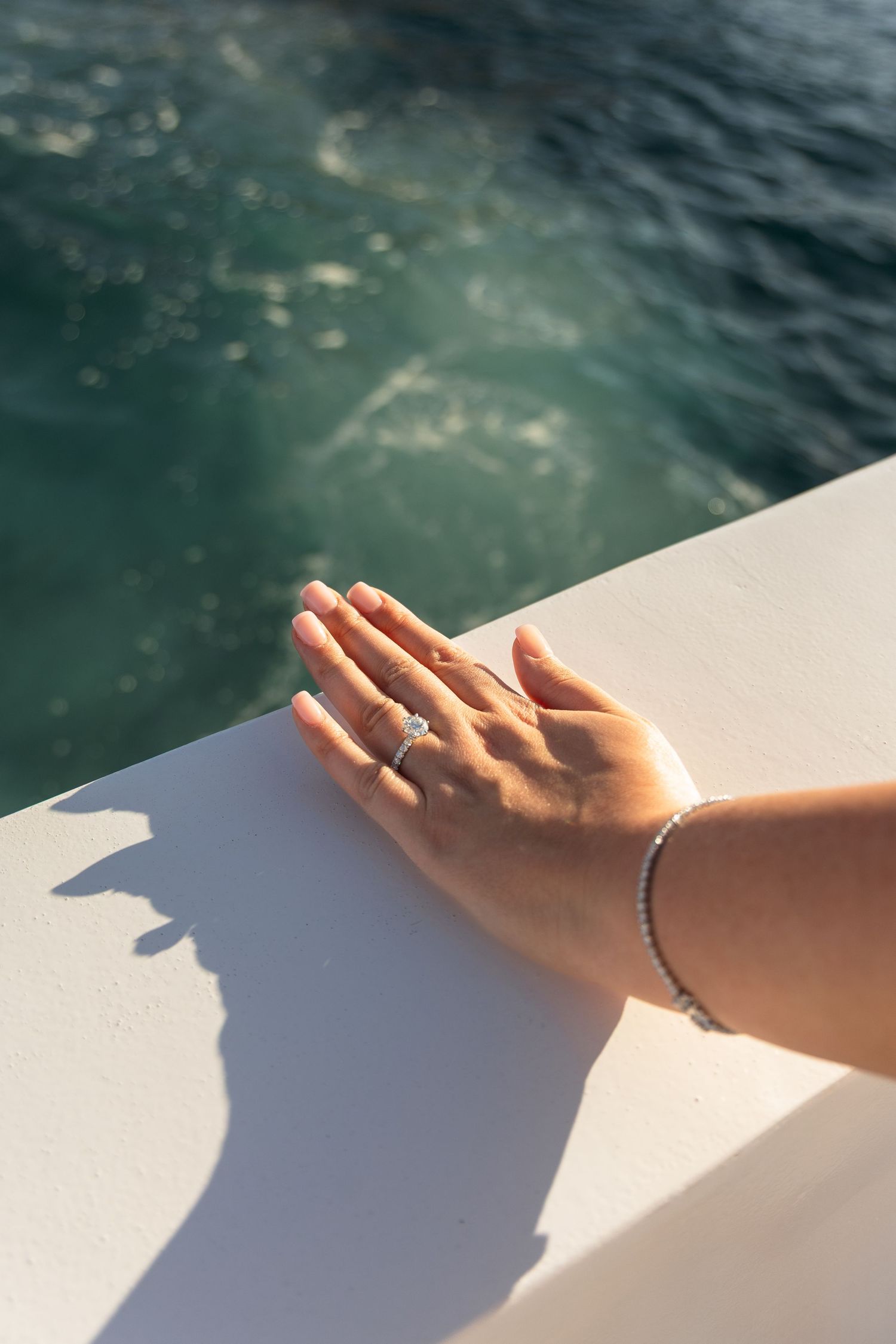 A delicate hand with a ring and bracelet rests on a white surface near turquoise water in sunlight.