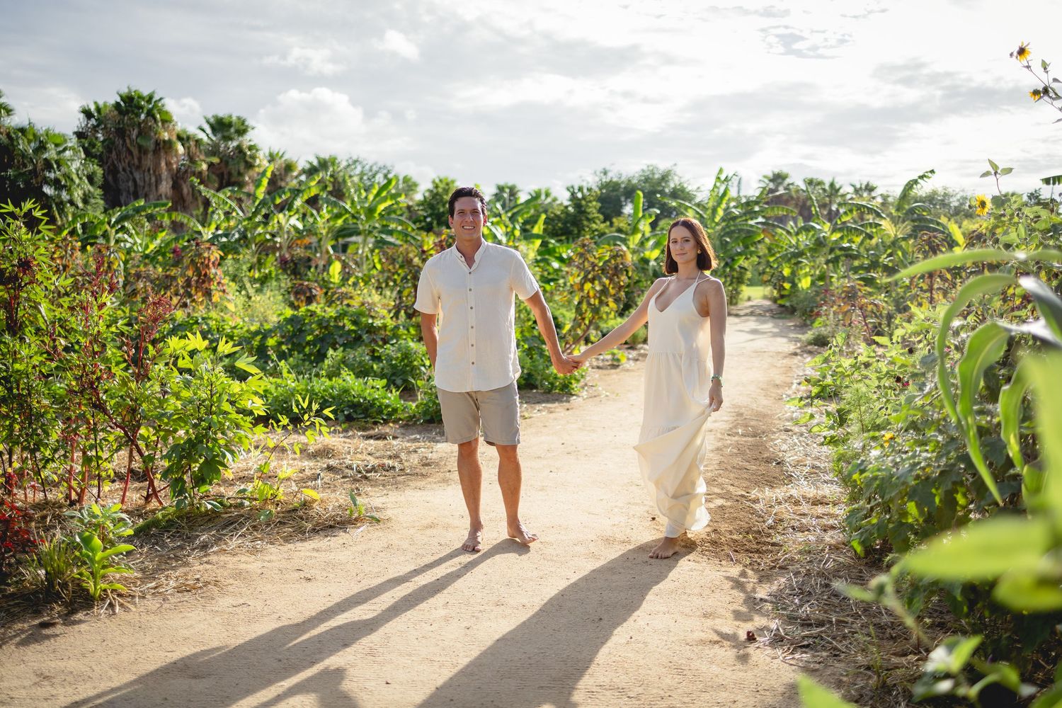 A couple walks hand in hand through a lush garden path surrounded by tropical foliage and flowers.
