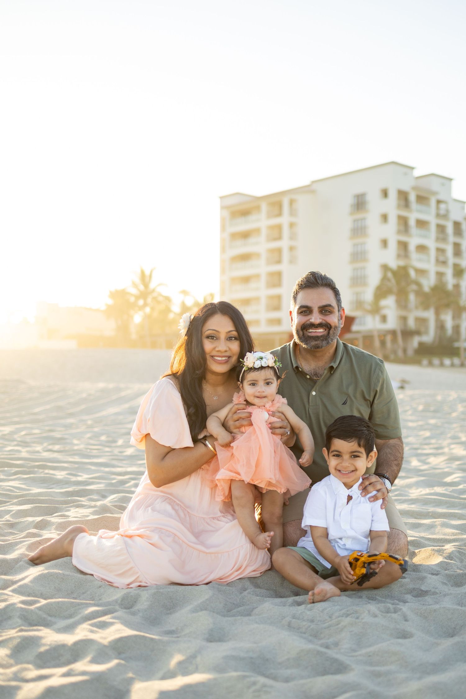 Family of four smiling on sandy beach at golden hour with resort building in background.