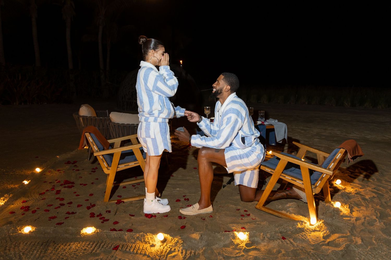 A romantic beach proposal scene at night with candles glowing in the sand around two wooden chairs.