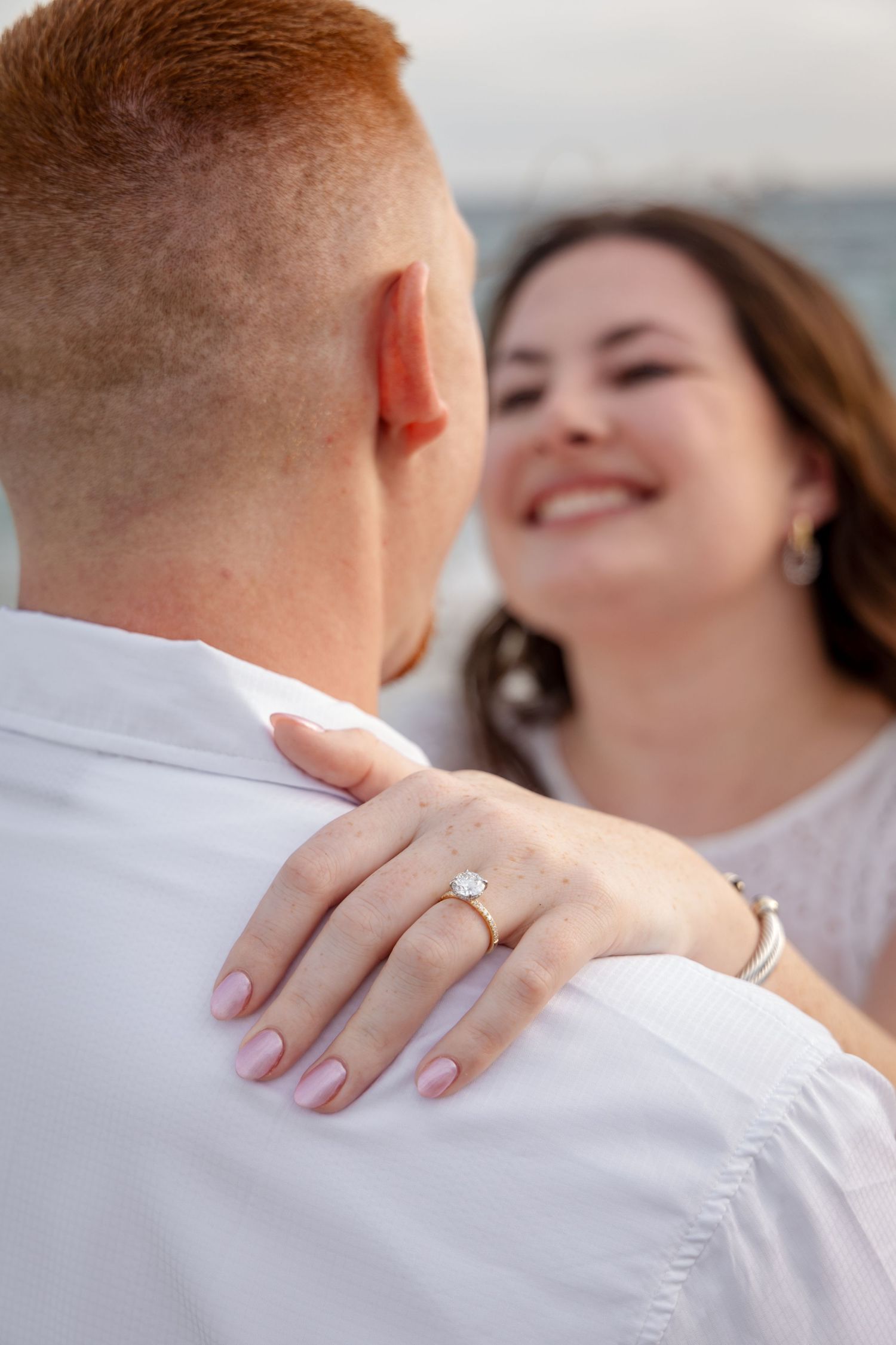 A couple embraces during an engagement photo session showing a close-up of hands and ring on white shirt.