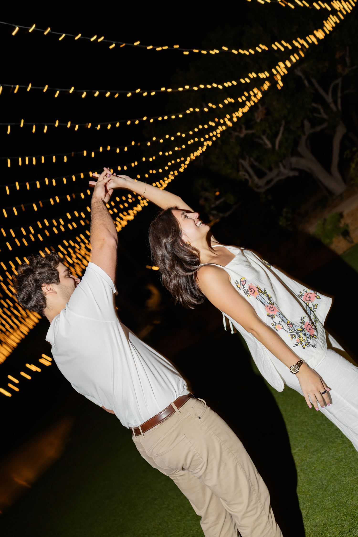 A couple dances and dips romantically under twinkling string lights at an outdoor evening celebration.