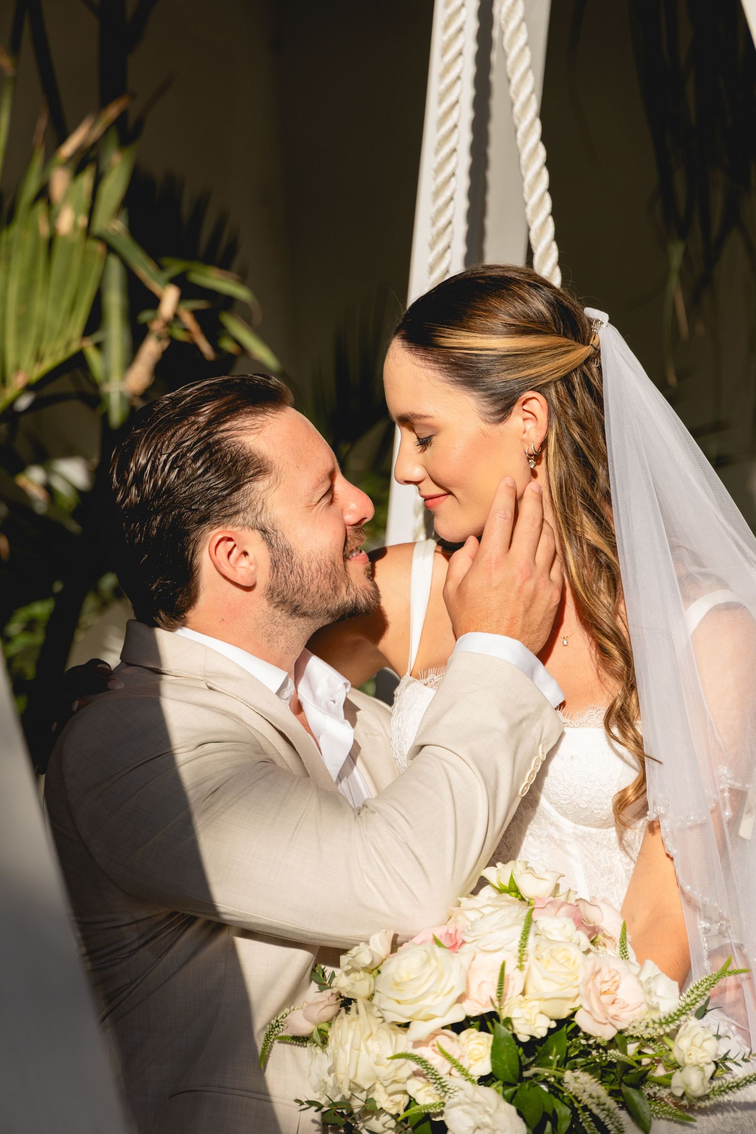 Romantic moment between newlyweds by white rope swing with floral bouquet.