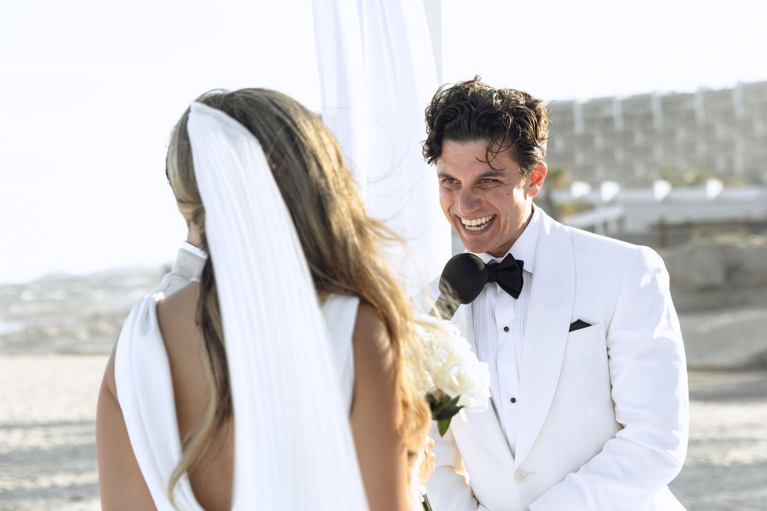 Candid moments between couple at oceanfront wedding ceremony under white canopy.