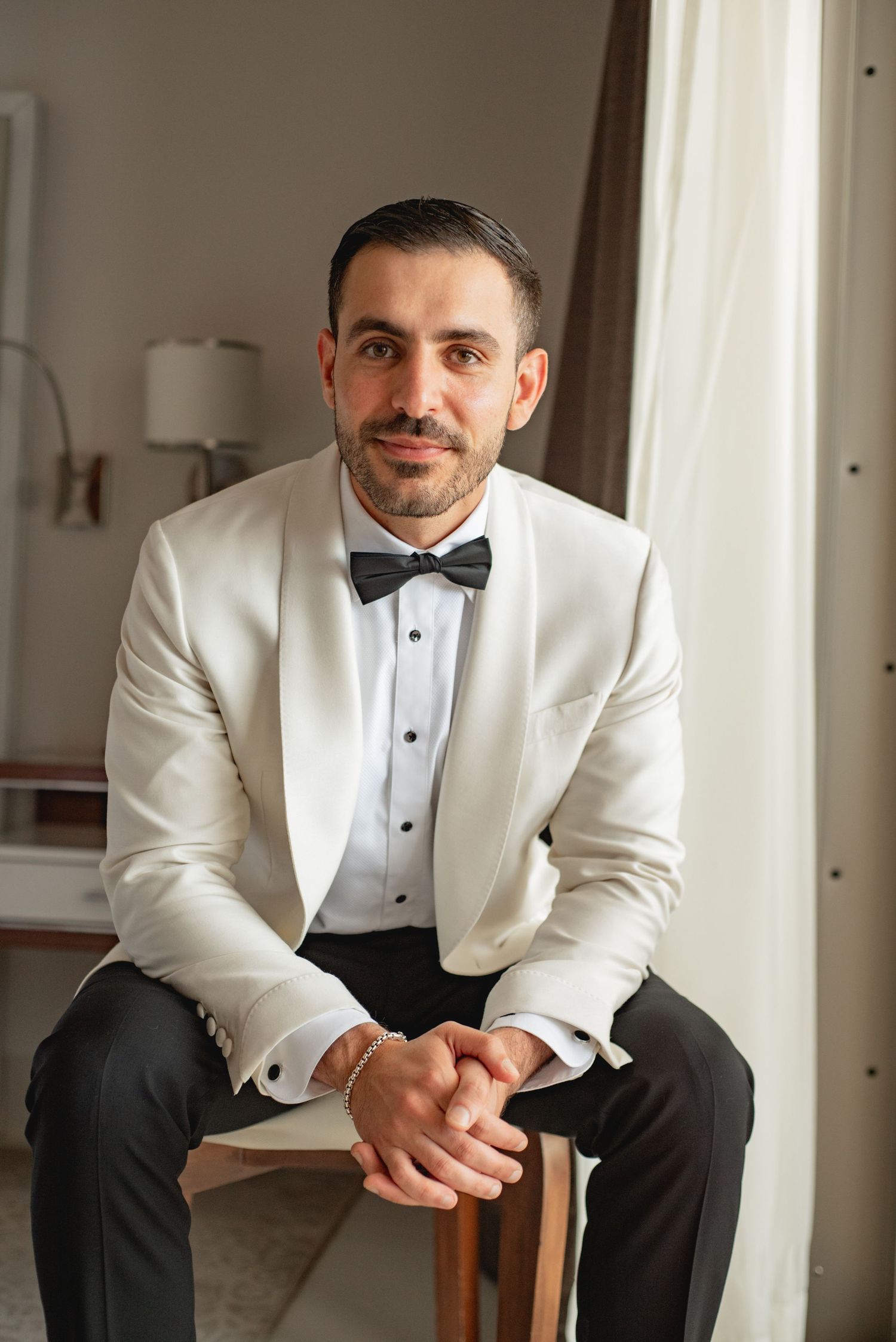 Groom in white tuxedo jacket and bow tie sitting on wooden chair for formal portraits.