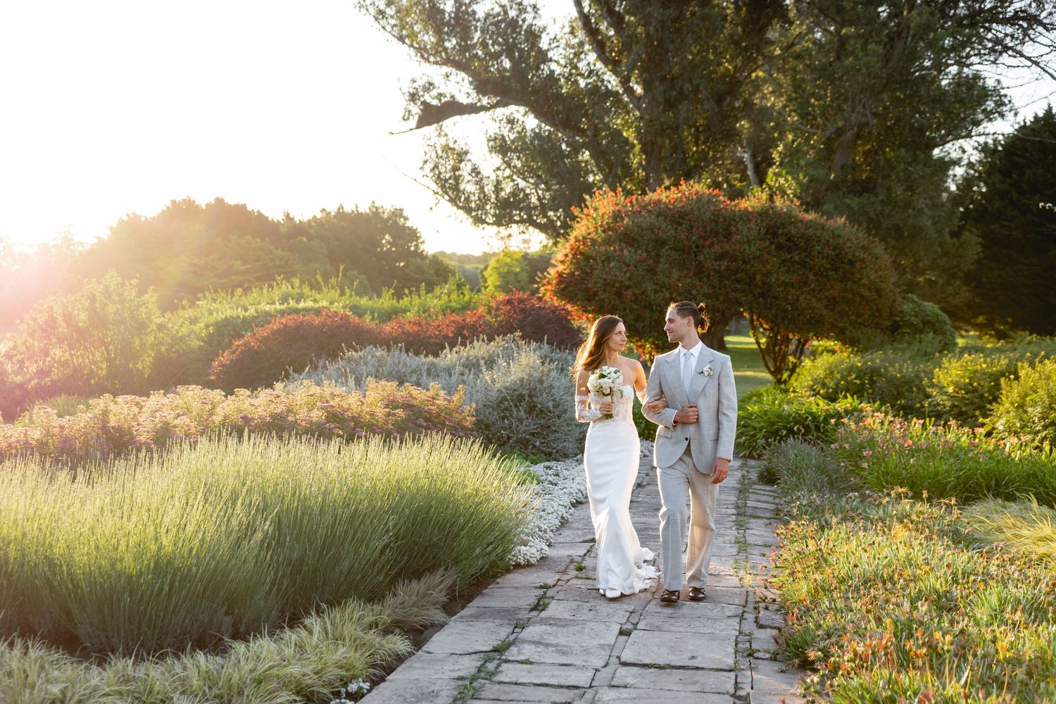 Wedding couple walking through a sunlit meadow with tall grass and trees.