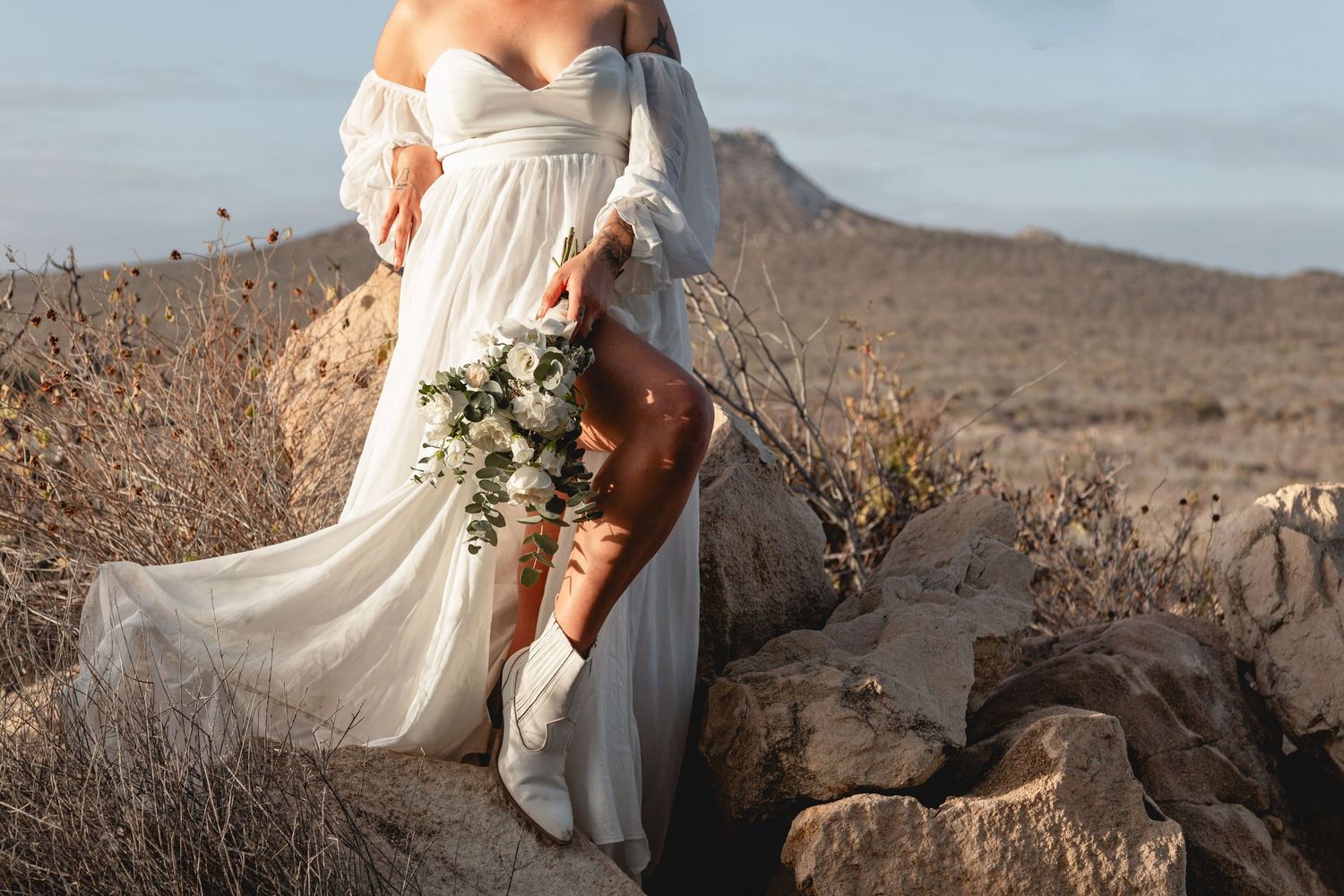 A flowing white wedding dress billows in the wind against a desert mountain landscape at sunset.