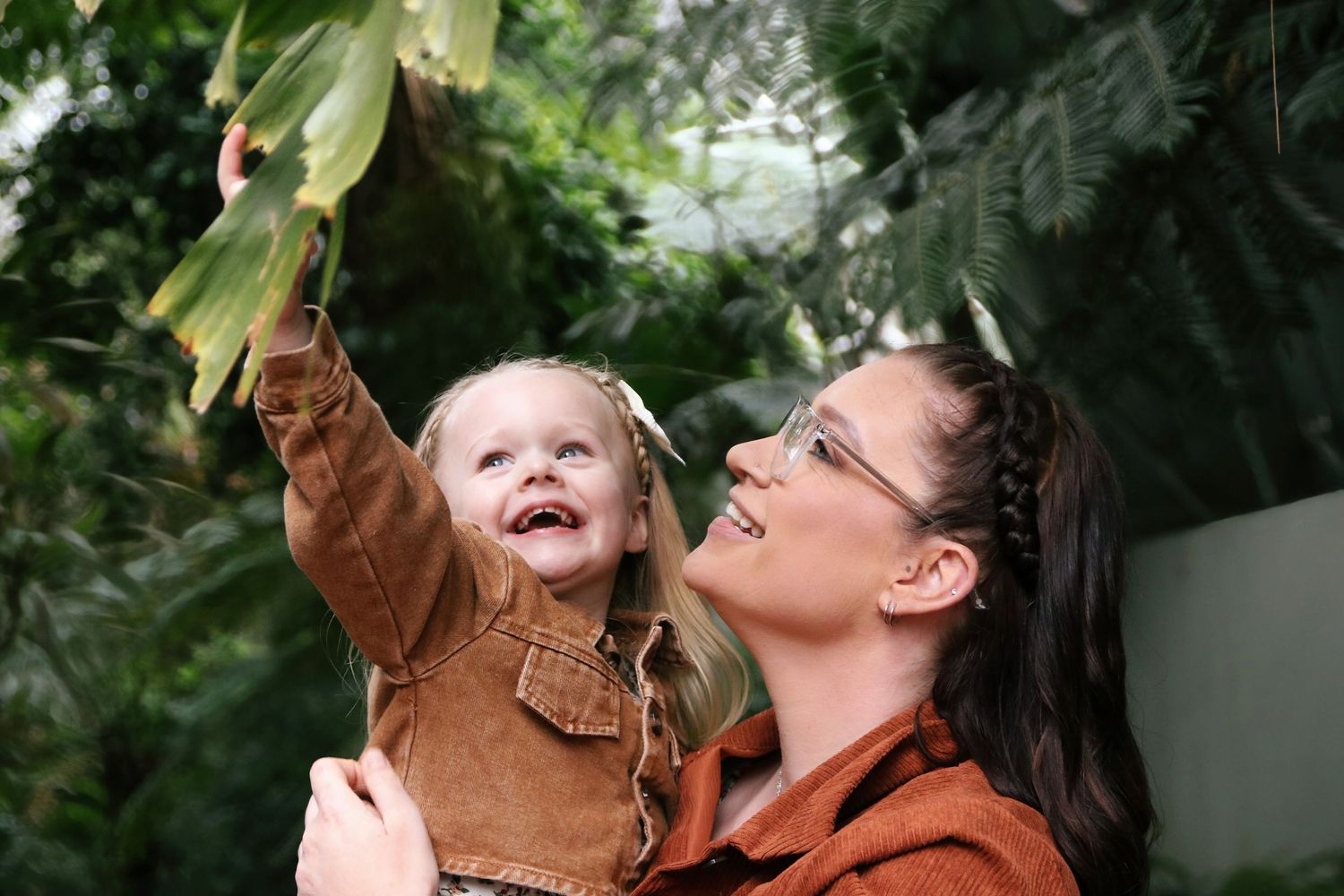 Woman holding smiling toddler girl reaching up to touch tropical green leaves in a lush greenhouse.
