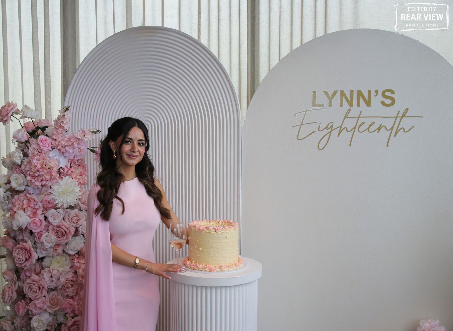 Young woman in pink dress poses with birthday cake at Lynn's Eighteenth birthday celebration with floral backdrop.