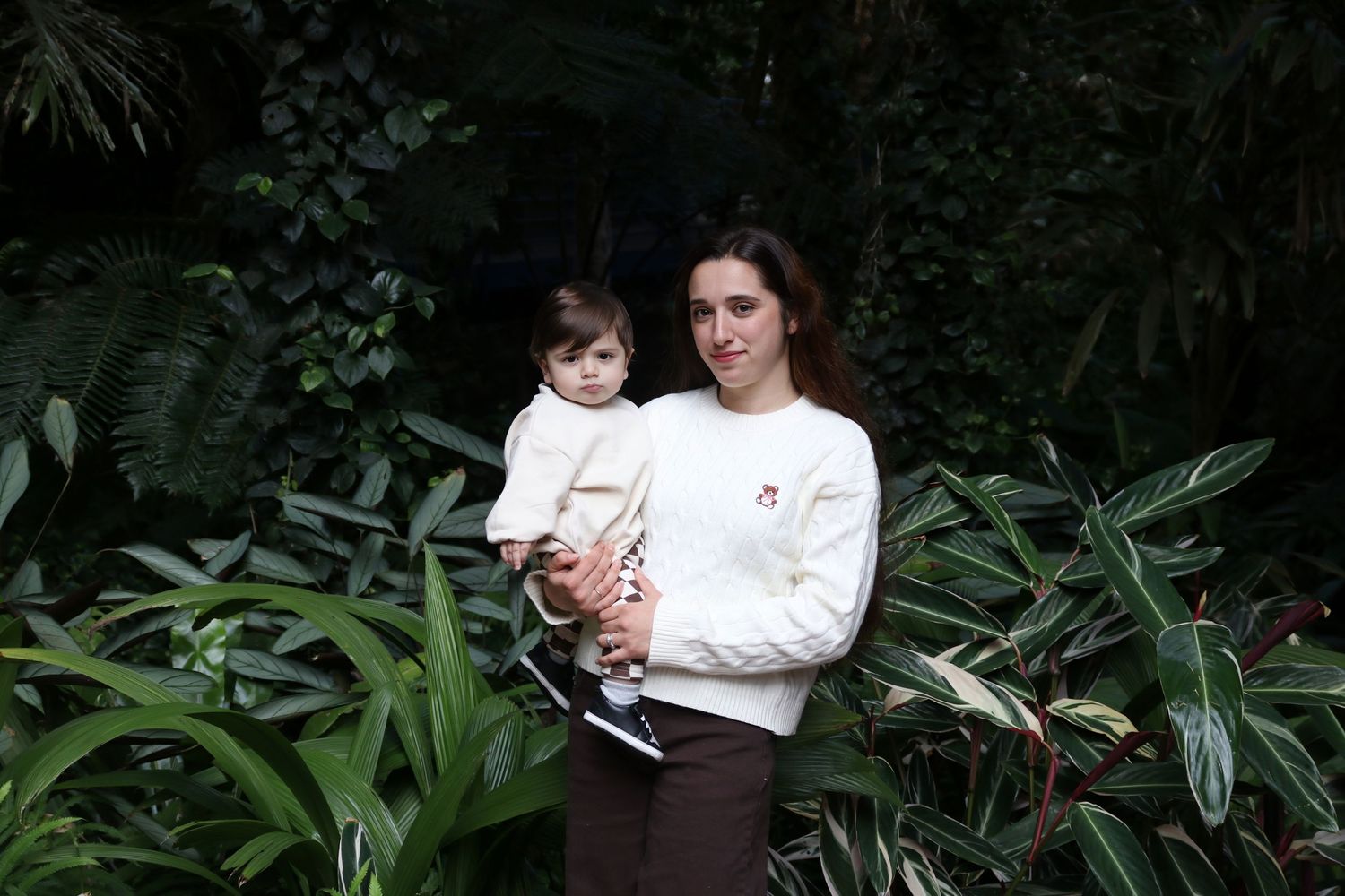Woman in white sweater holding a baby amid lush tropical green foliage outdoors.