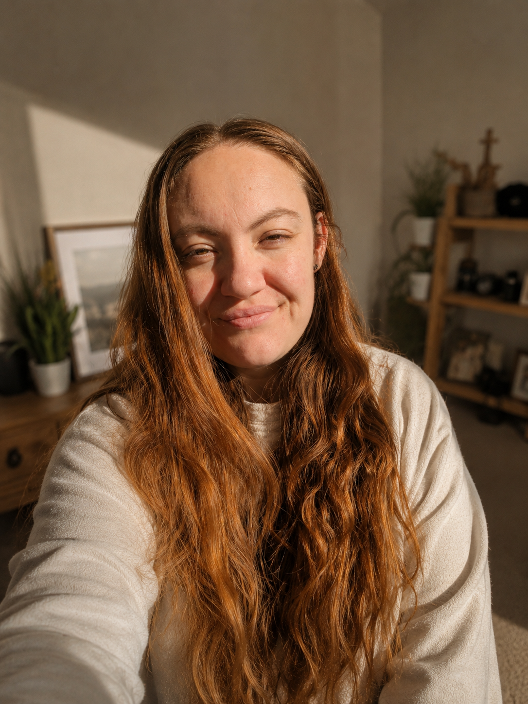 A smiling woman with long auburn wavy hair wearing a white sweater takes a selfie in a warmly lit home interior.