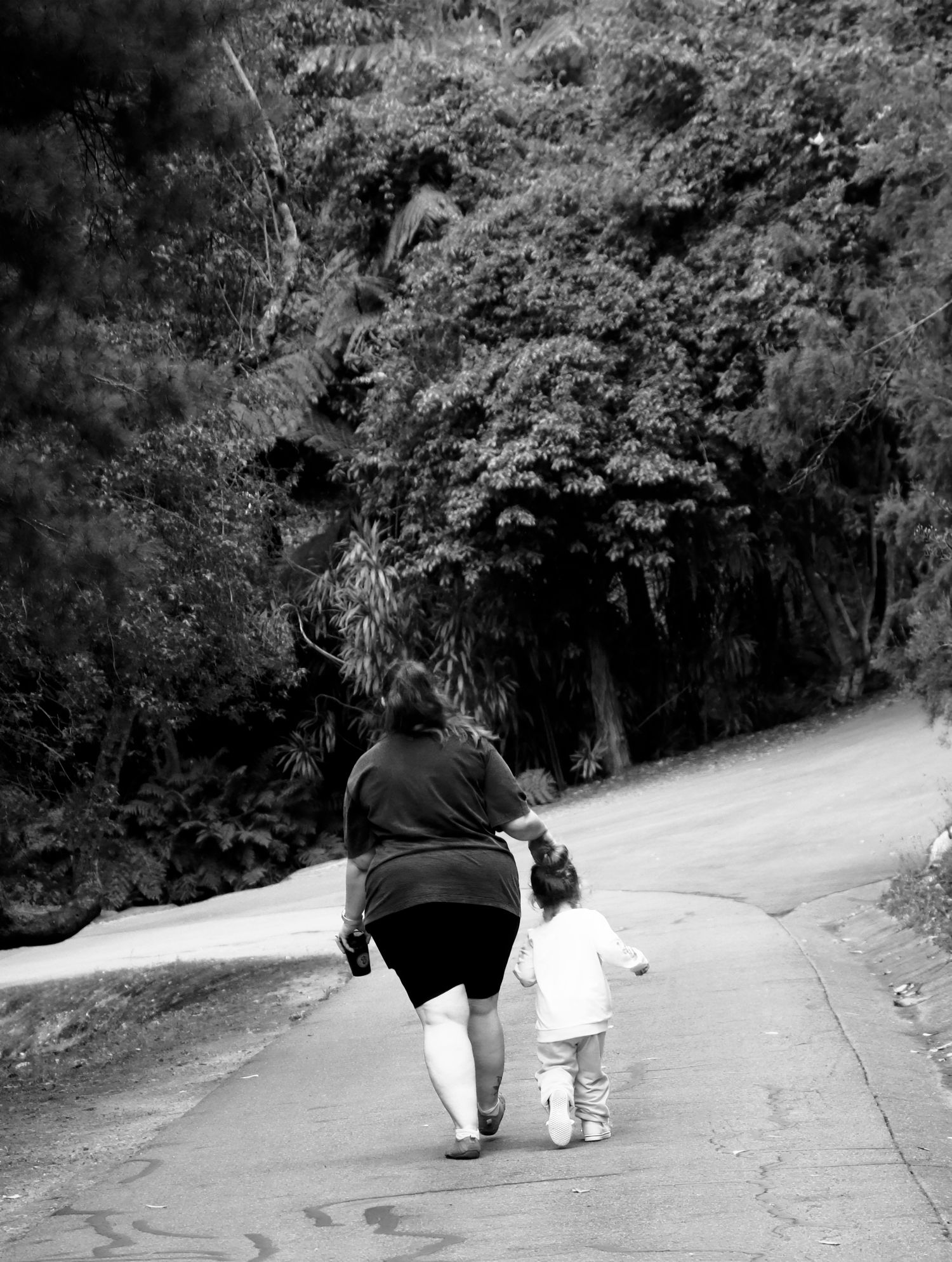 Black and white photo of a mother and young child walking hand-in-hand down a tree-lined road.