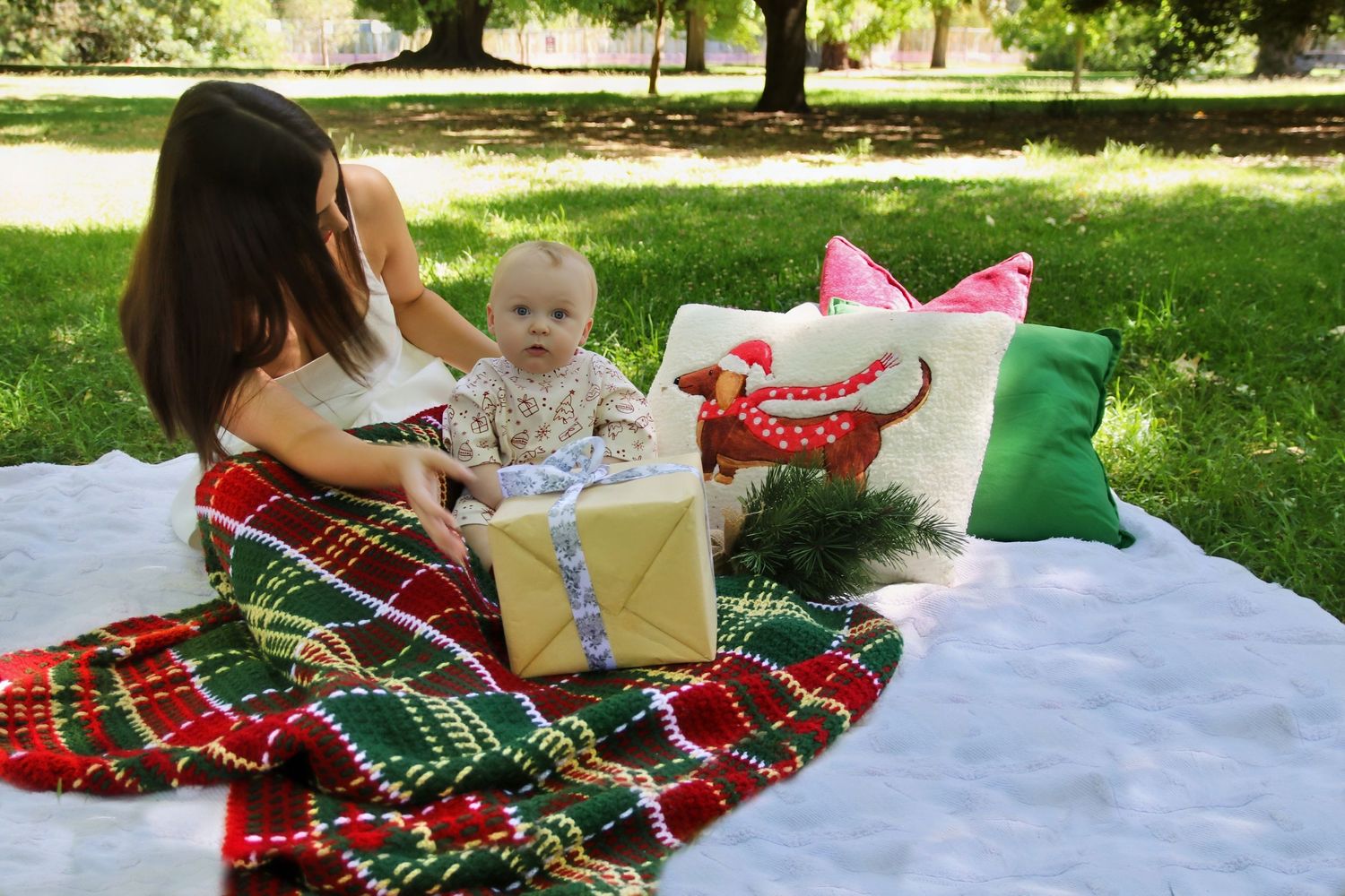 Mother and baby enjoying a Christmas picnic outdoors on a plaid blanket with holiday pillows and a gift.