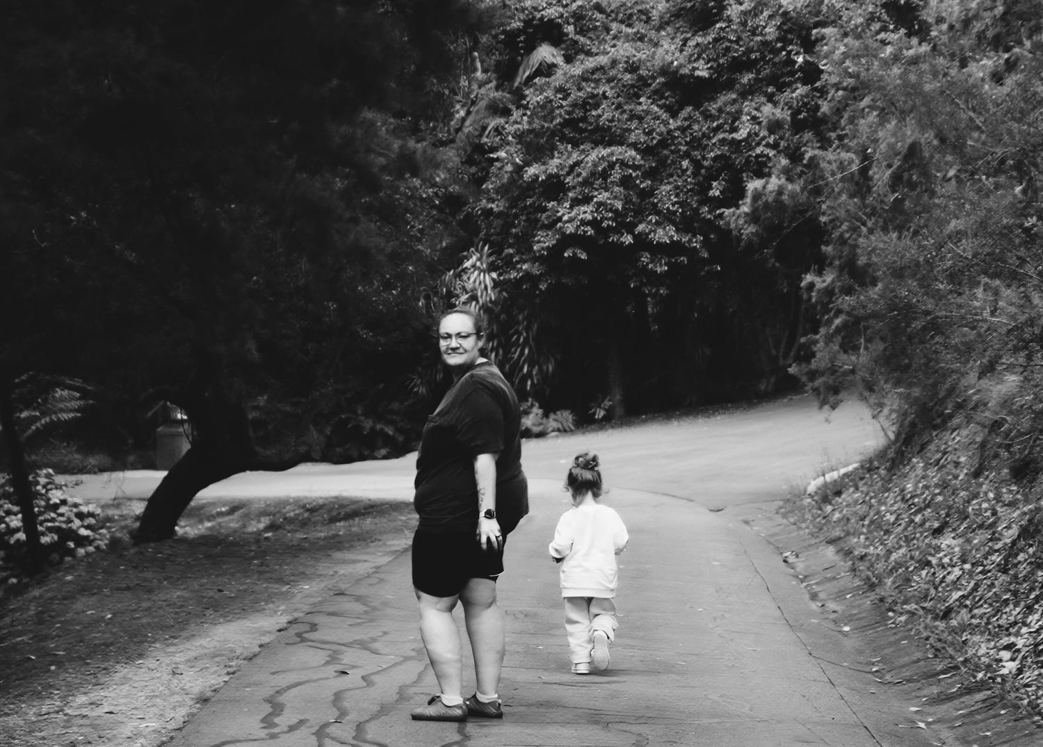 Black and white photo of a woman and young child walking hand in hand on a tree-lined road.