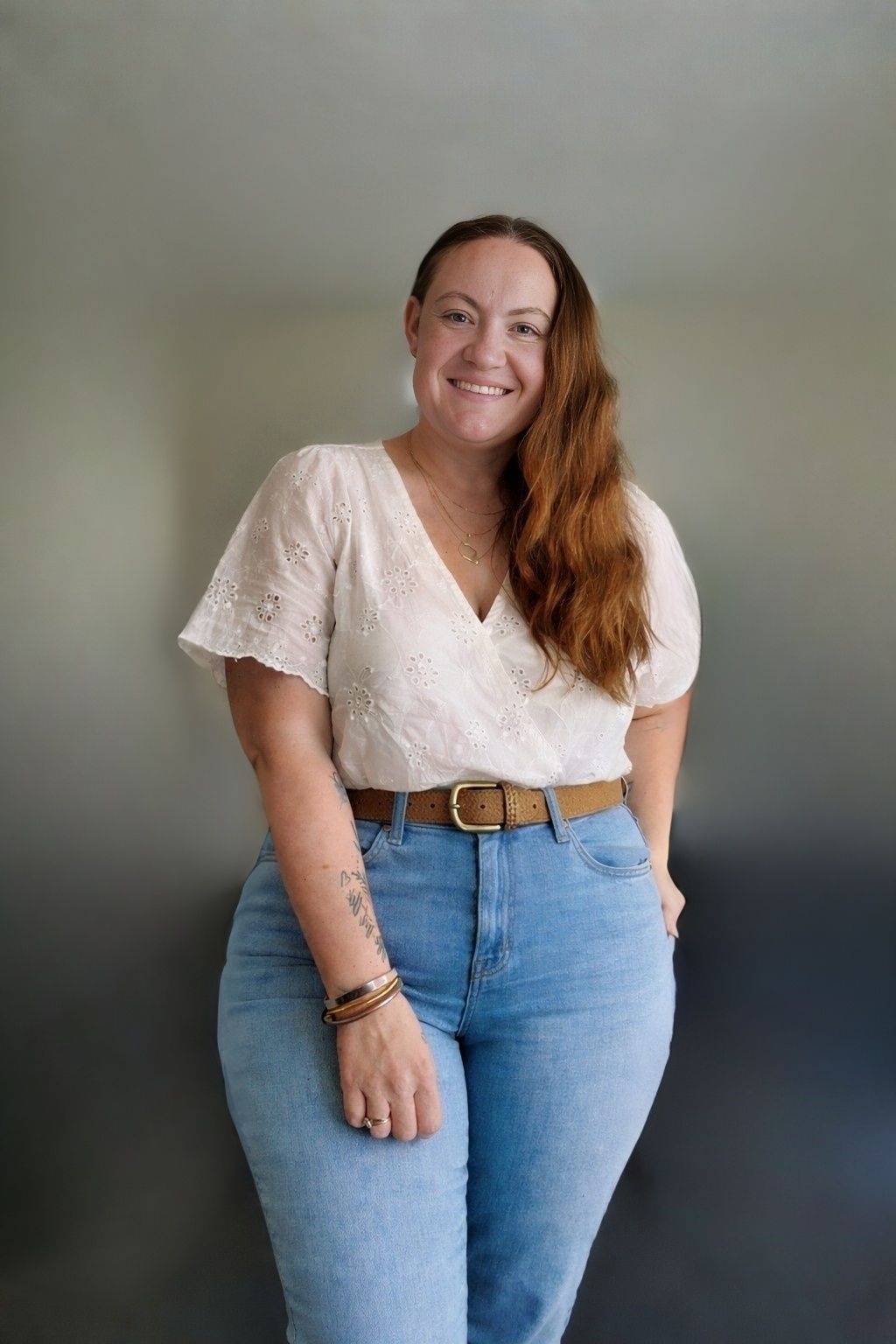 Smiling woman with long auburn hair wearing a white eyelet top, brown belt, and light blue jeans poses indoors.
