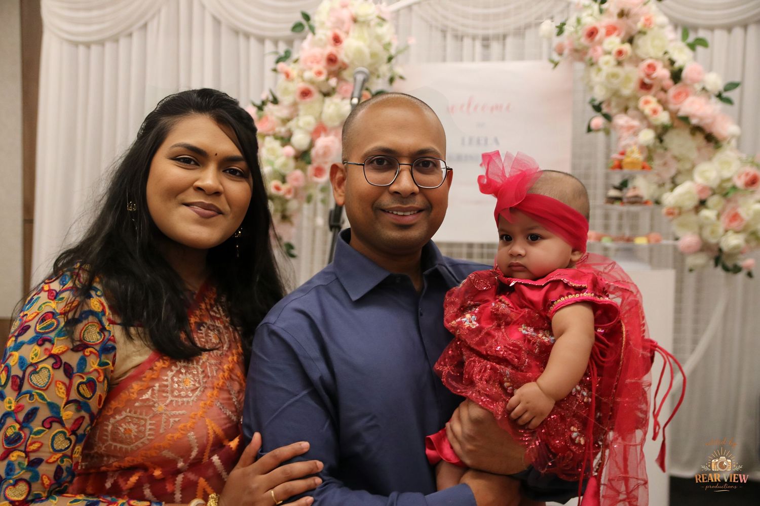 A South Asian family poses with their baby girl in a red dress at a floral decorated celebration event.