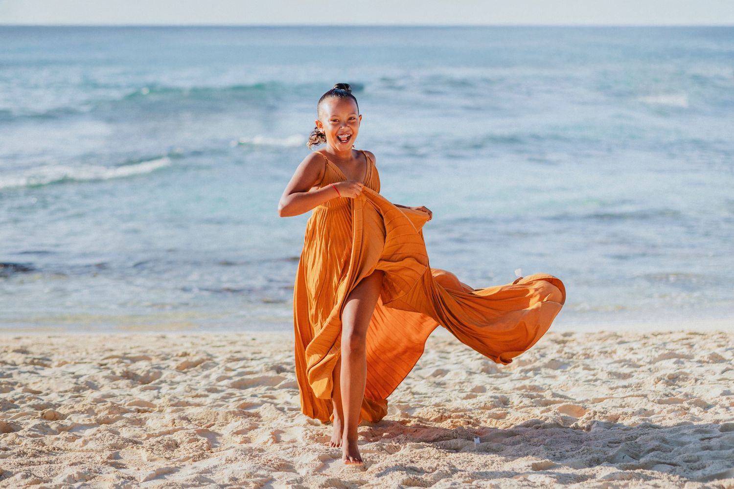 Child in flowing orange dress twirls and plays on sandy beach.
