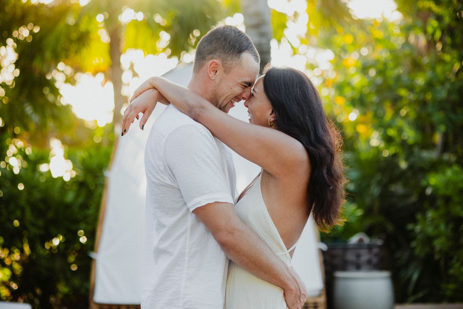 Romantic couple in white clothing embracing in sunlit tropical garden setting.