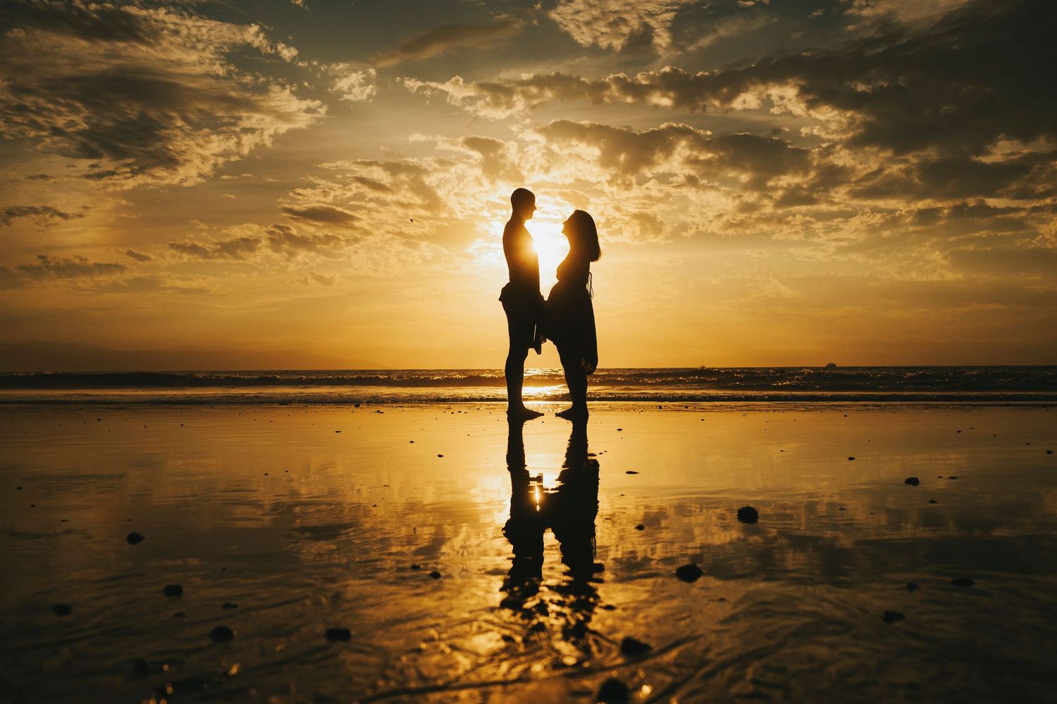 Silhouettes of figures walking along a beach at sunset with dramatic golden clouds reflecting on wet sand.