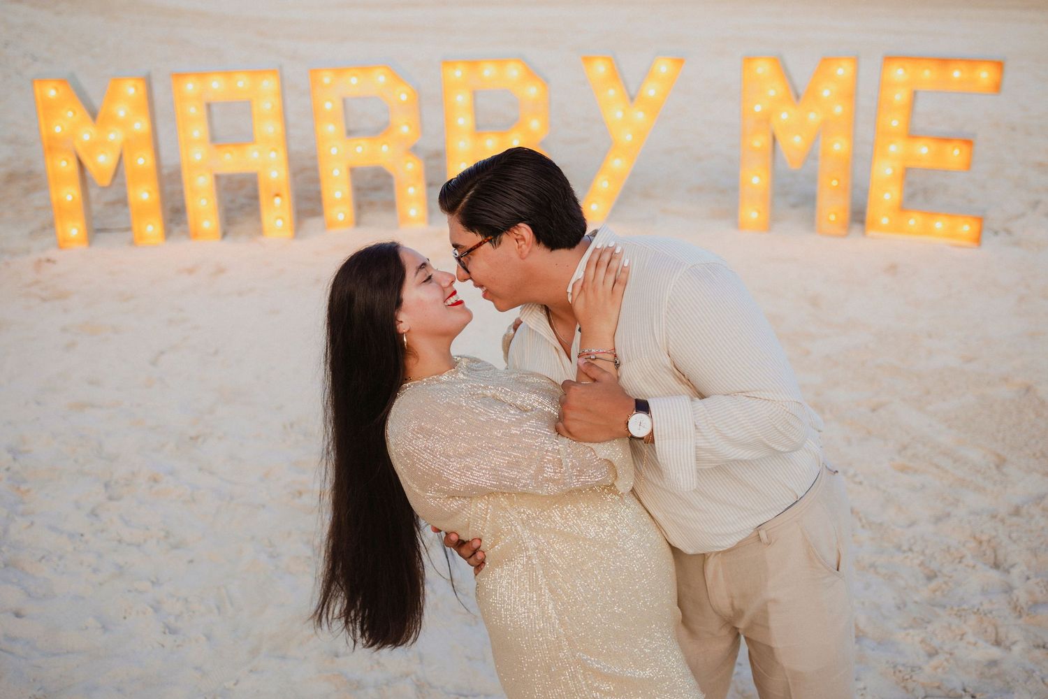 Beach proposal scene with illuminated 'Marry Me' text written in the sand at dusk.