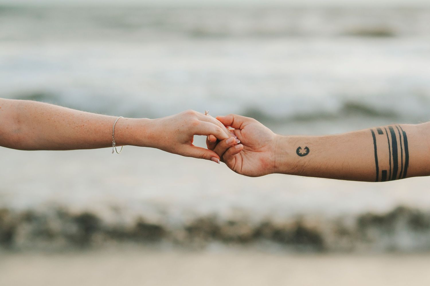 Close-up of tattooed hands reaching toward each other against beach background.
