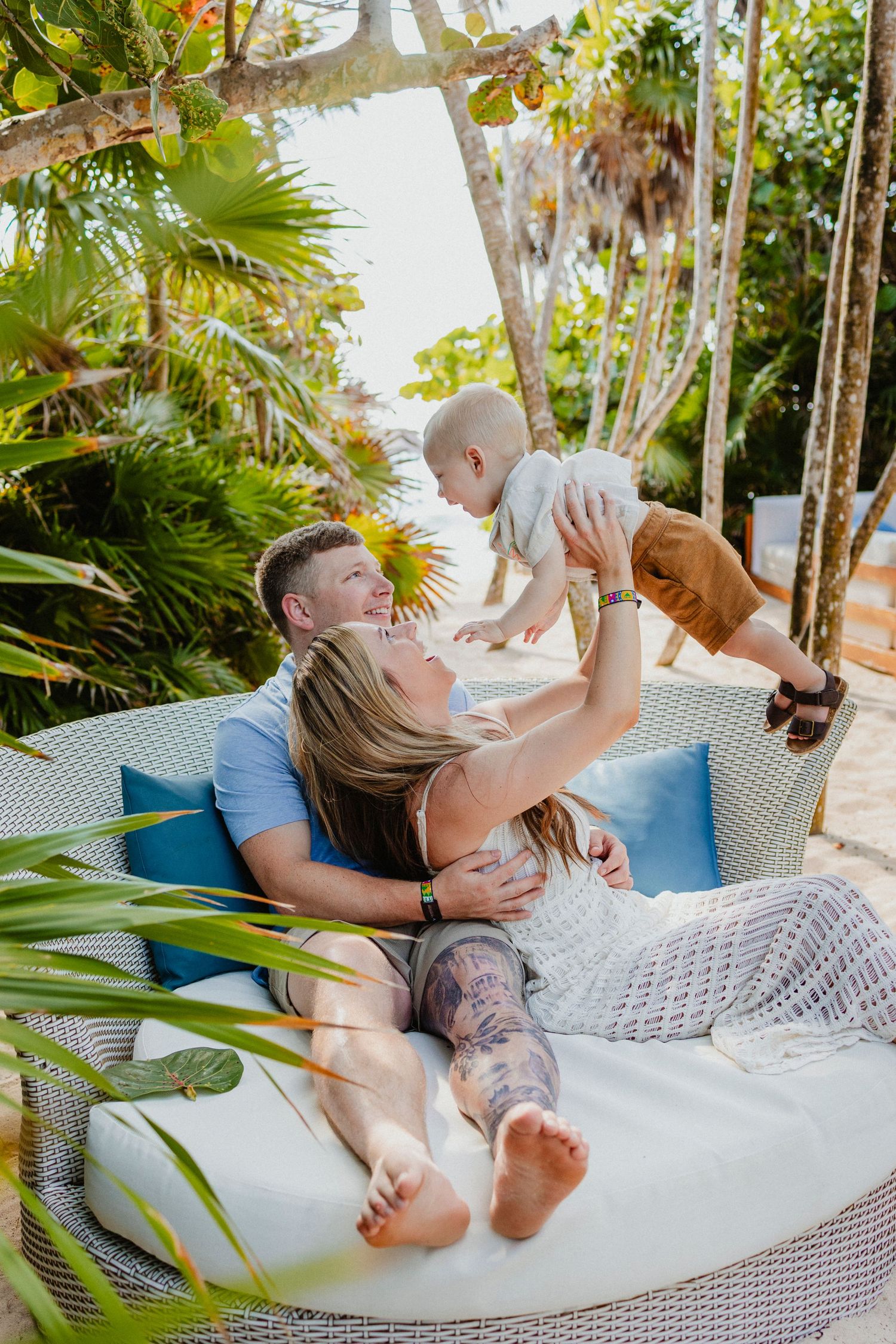 Family playing together on outdoor wicker furniture surrounded by tropical plants.