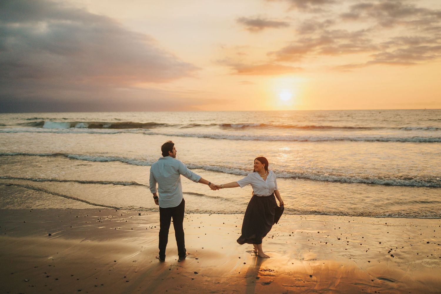 Couple walking along a scenic beach at sunset with waves crashing on the shore.