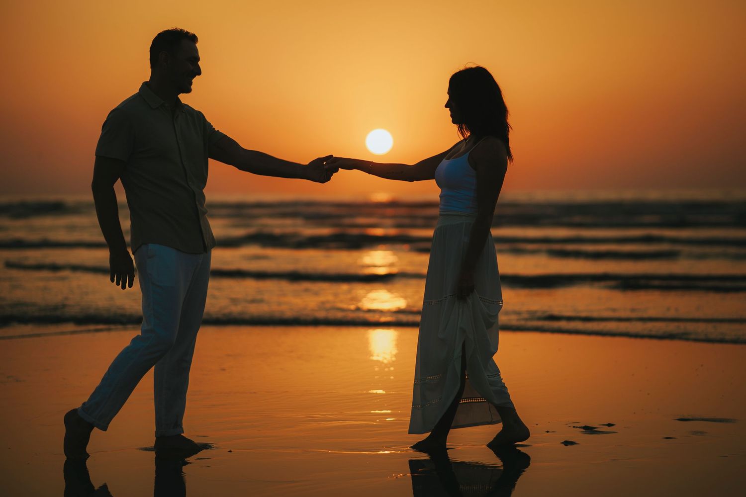 Silhouettes of people dancing and playing on beach at sunset with orange sky reflection.