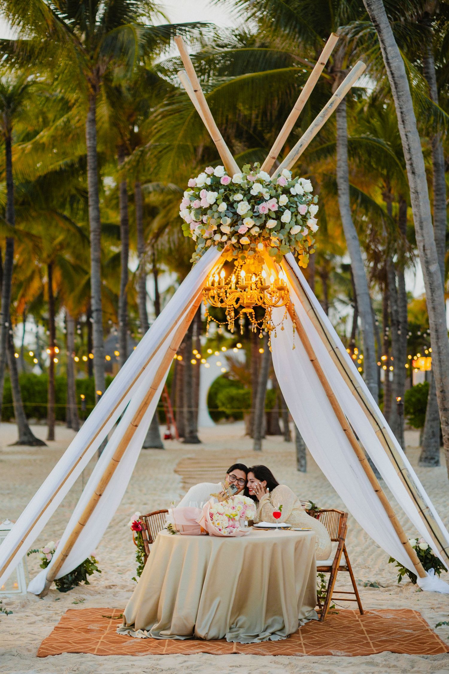 Elegant beach reception setup with white teepee tents, floral arrangements and string lights among palm trees.