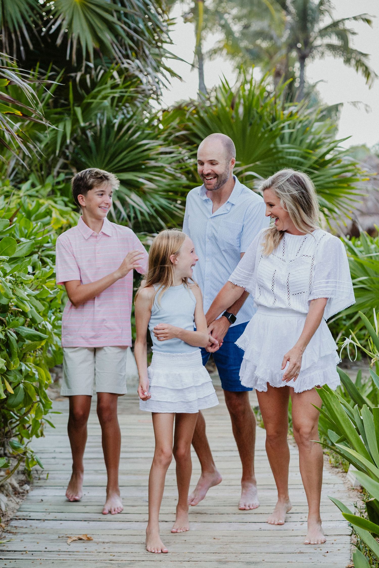 Family walks barefoot along tropical boardwalk surrounded by palm trees.