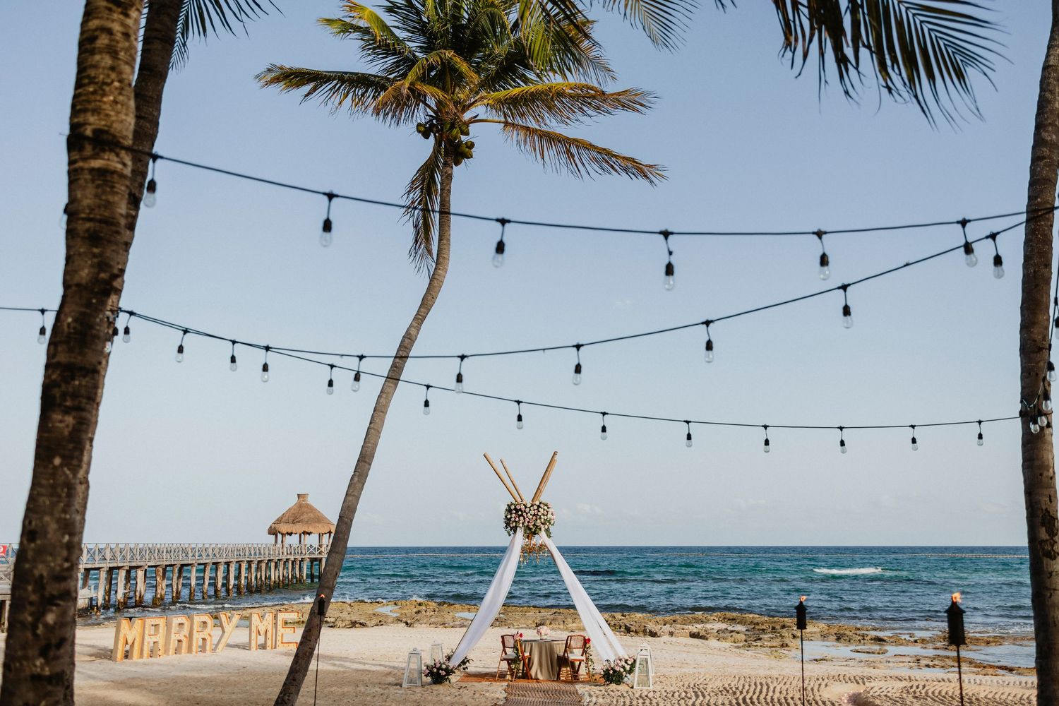 Tropical beach setting with string lights hanging between palm trees and tiki huts along the shore.